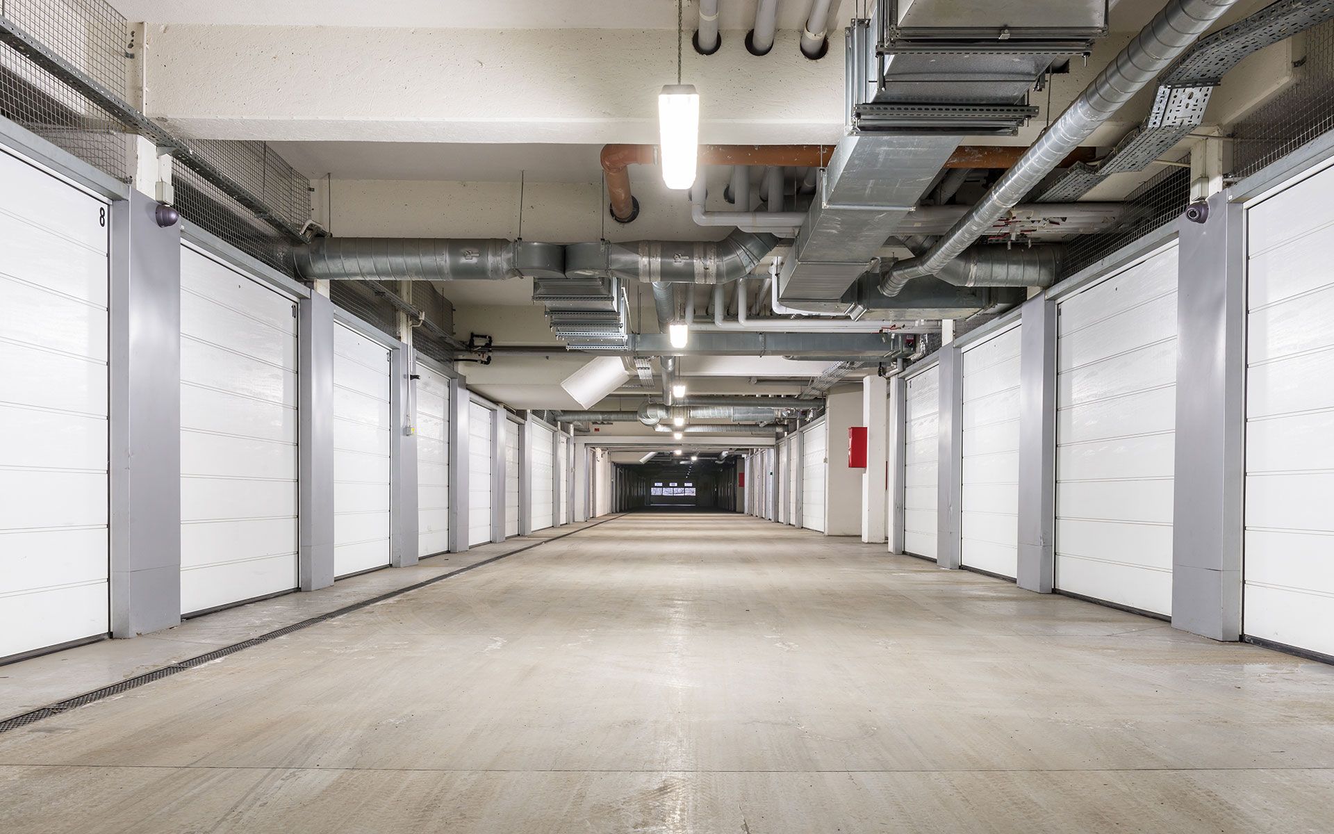 Long, concrete hallway with white storage units on both sides, overhead pipes and lights.