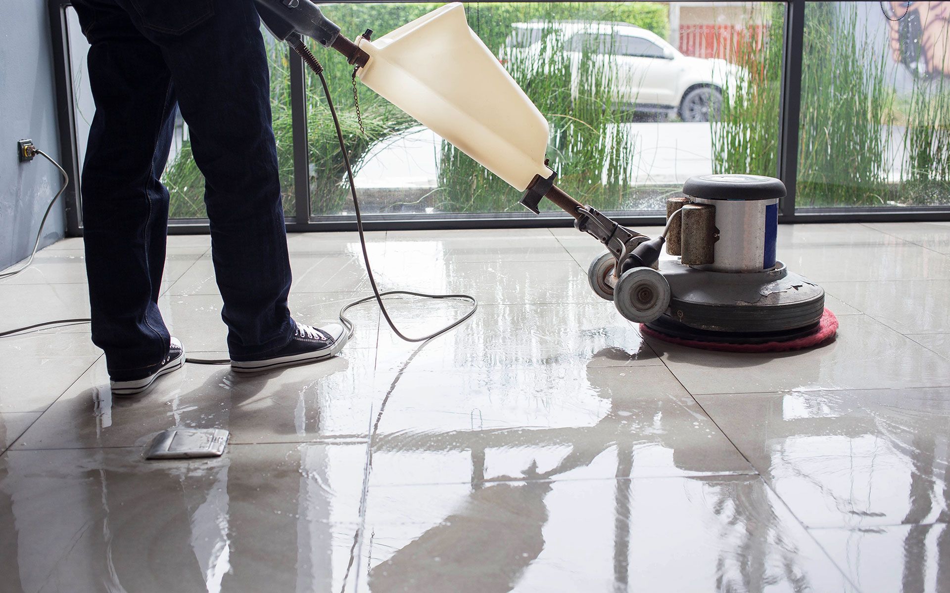 Person using a floor buffer on a wet, shiny tile floor inside a building near a window.