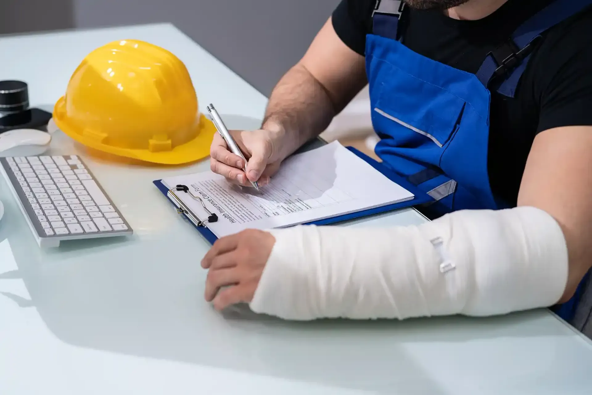 Person with bandaged arm filling out paperwork at a desk, yellow hard hat nearby.