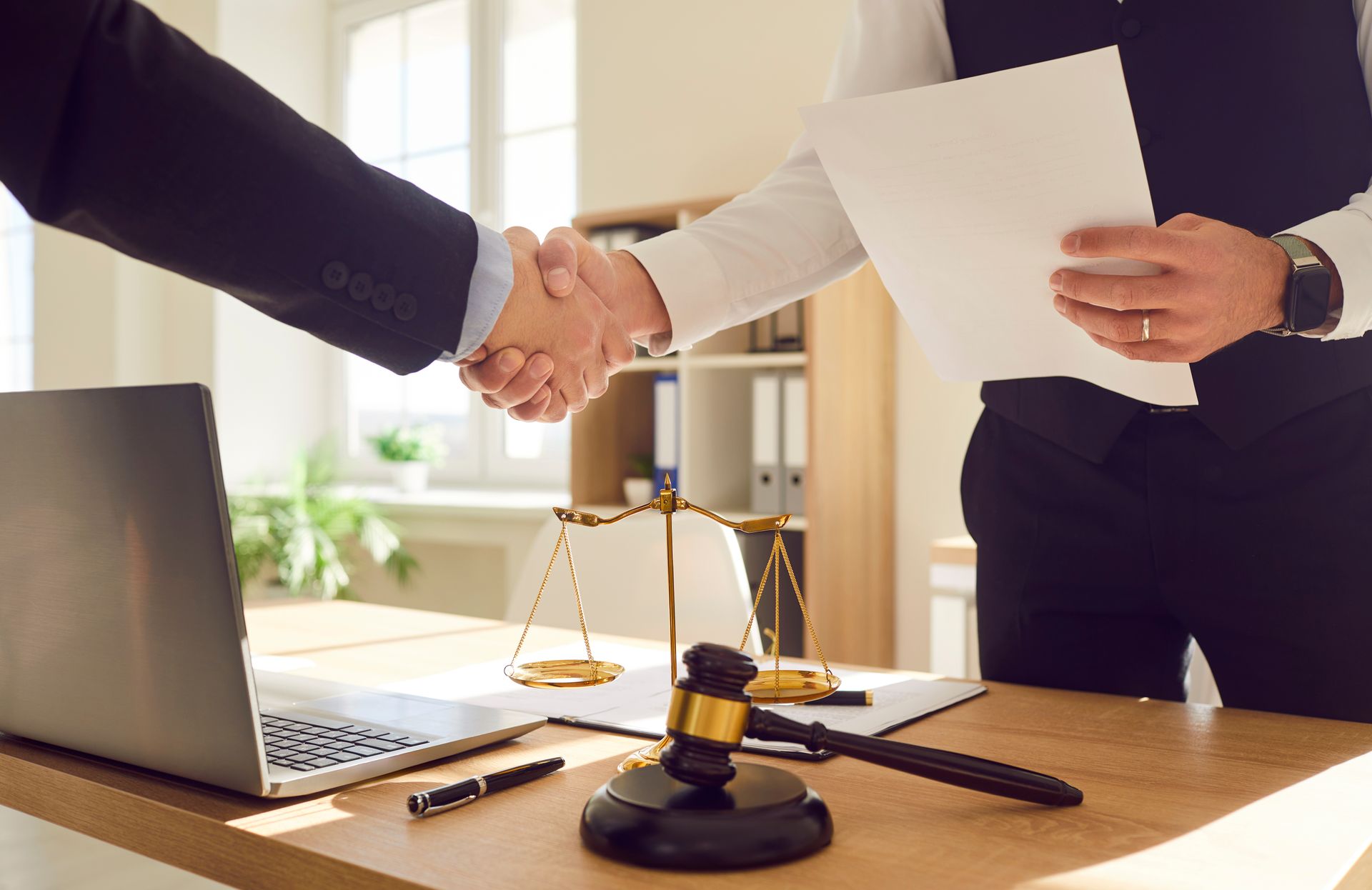 Two people shaking hands over a desk with a gavel, scales, and laptop, in a law office.