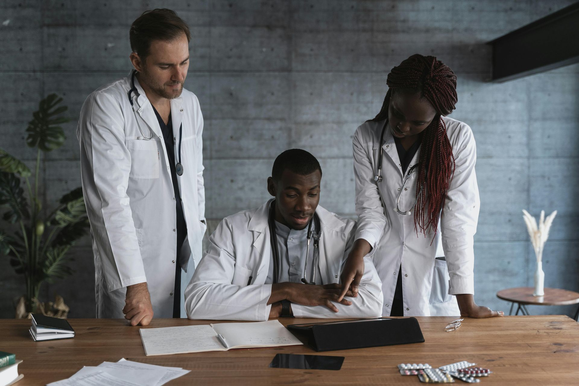 Doctors in white coats examining documents at a wooden table in a room with a concrete wall.
