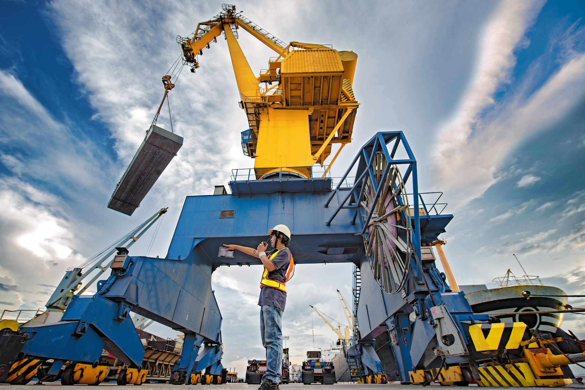 Trabajador dirigiendo una gran grúa amarilla que levanta metal en un puerto bajo un cielo parcialmente nublado.