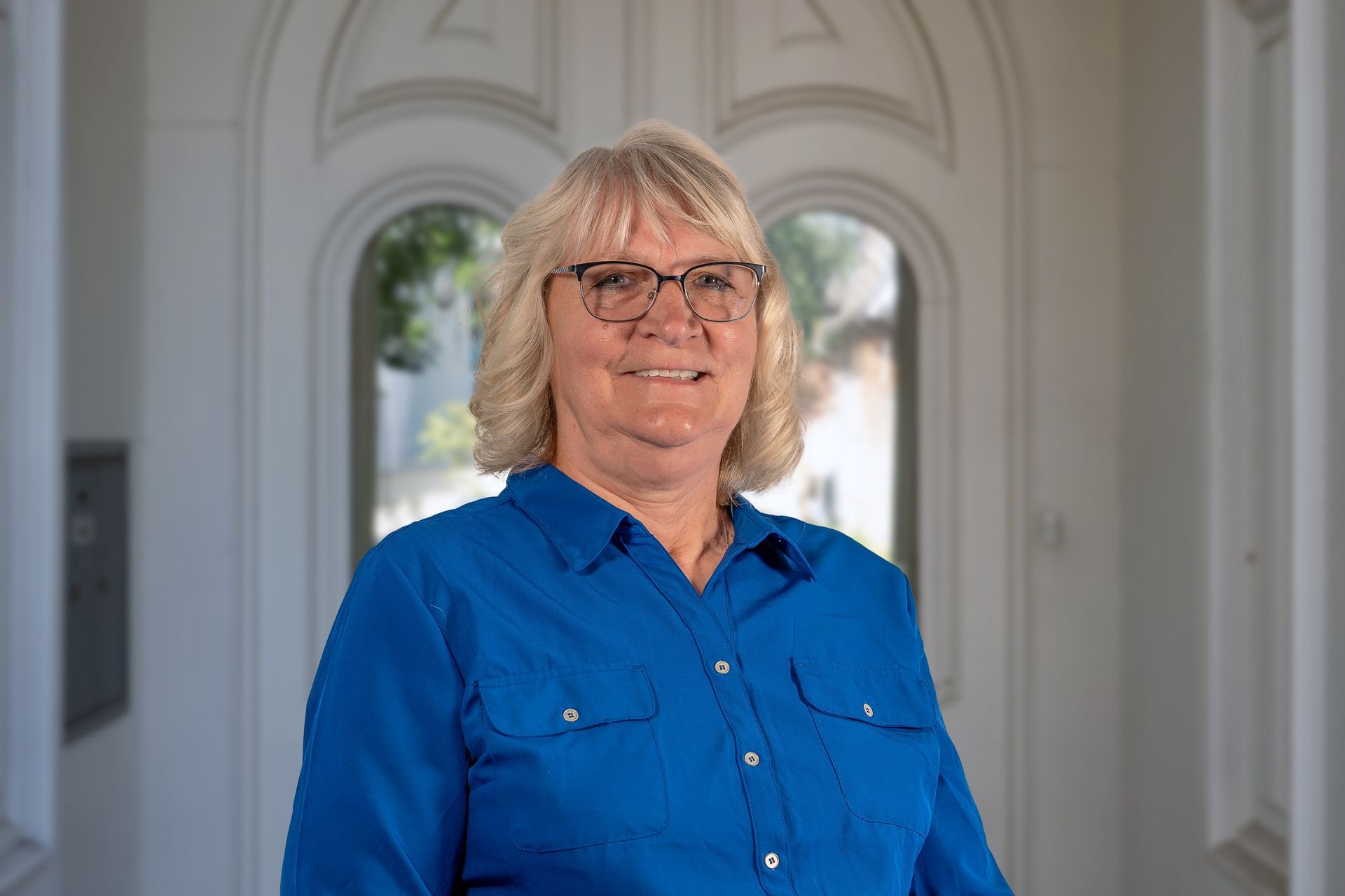 Woman in blue shirt and glasses smiles in a bright hallway.