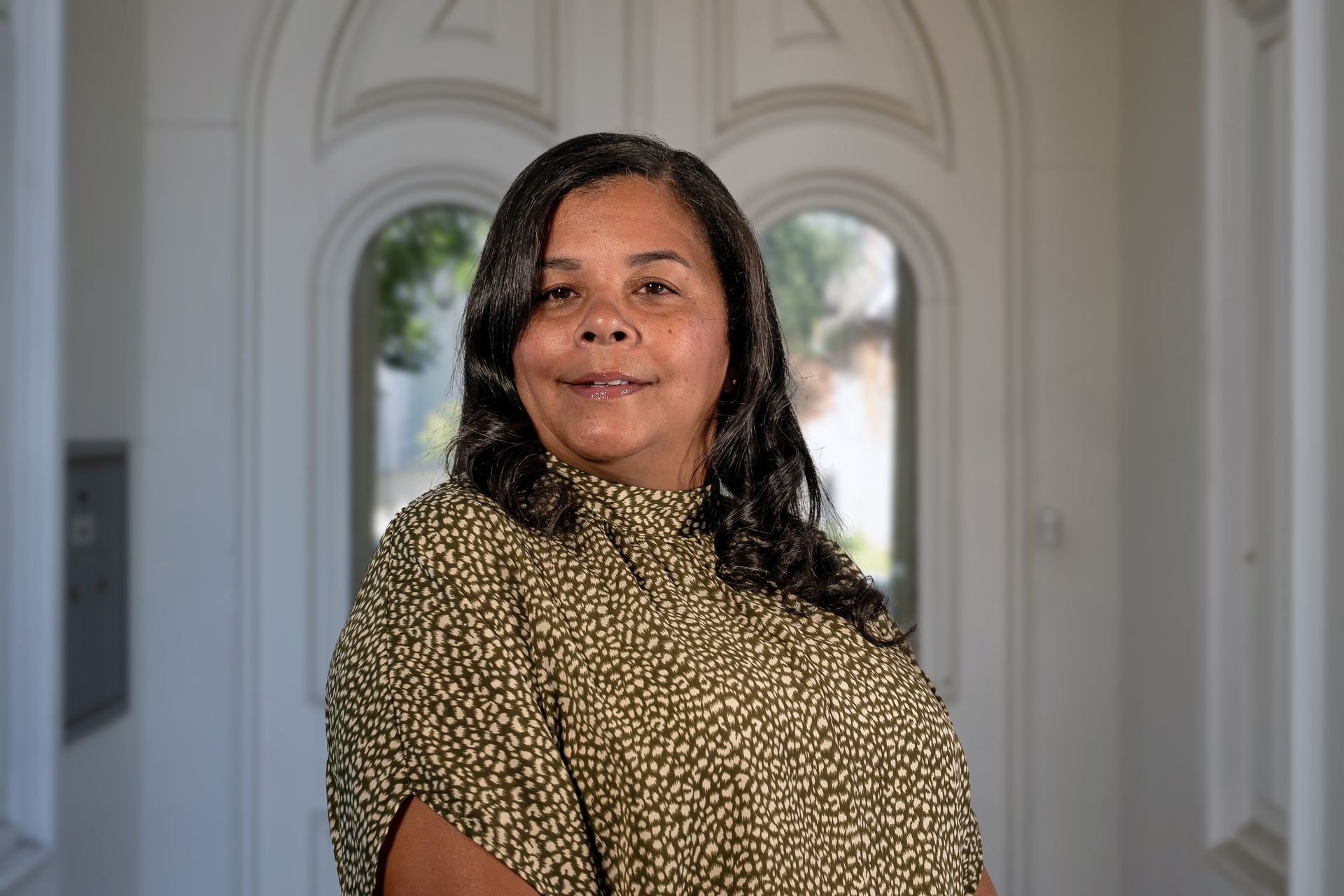 Woman in olive-colored patterned top smiles, standing in front of a white doorway.