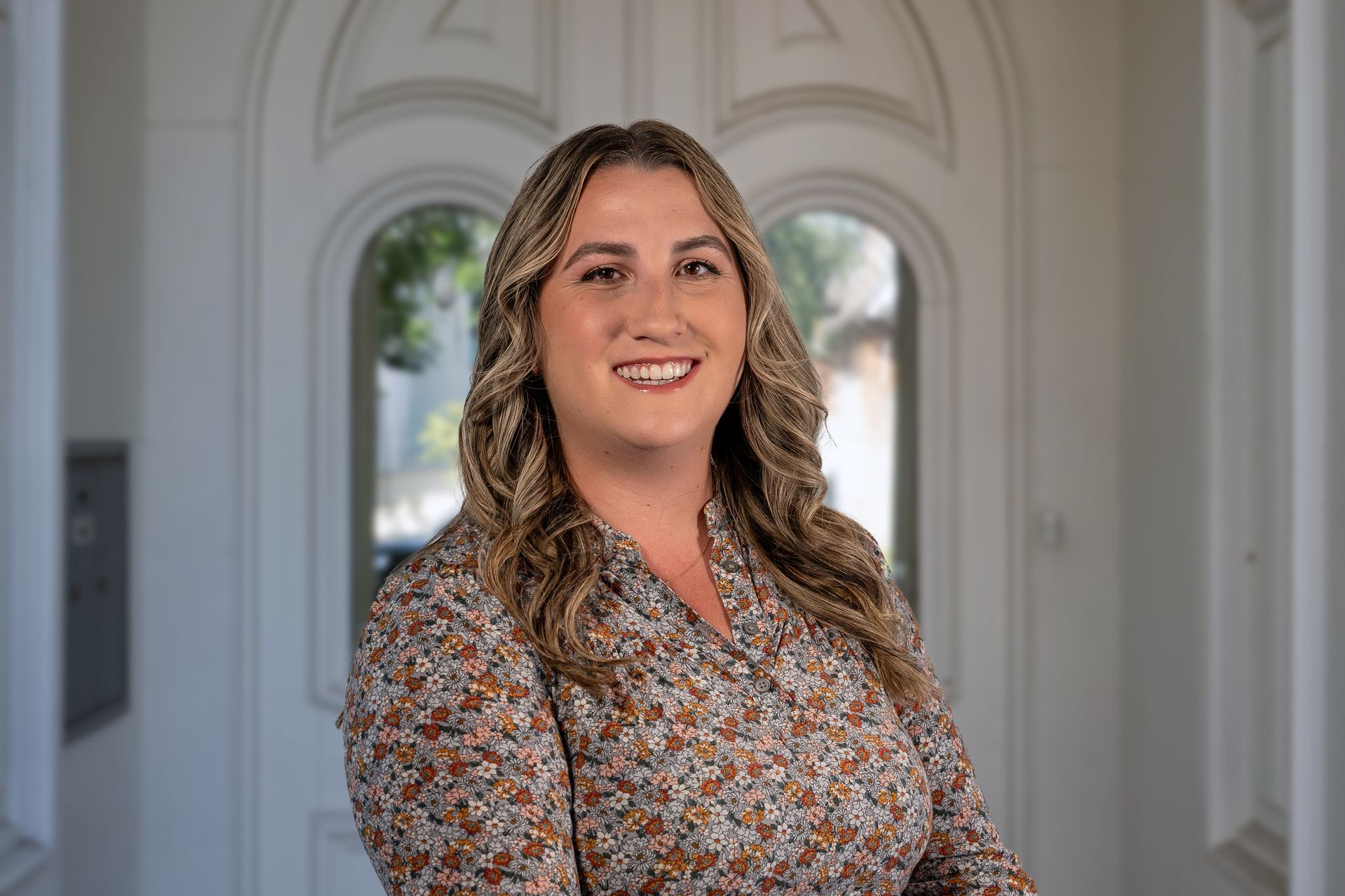 Woman smiles, stands in front of a white, ornate door. She wears a floral print top, has long, wavy hair.