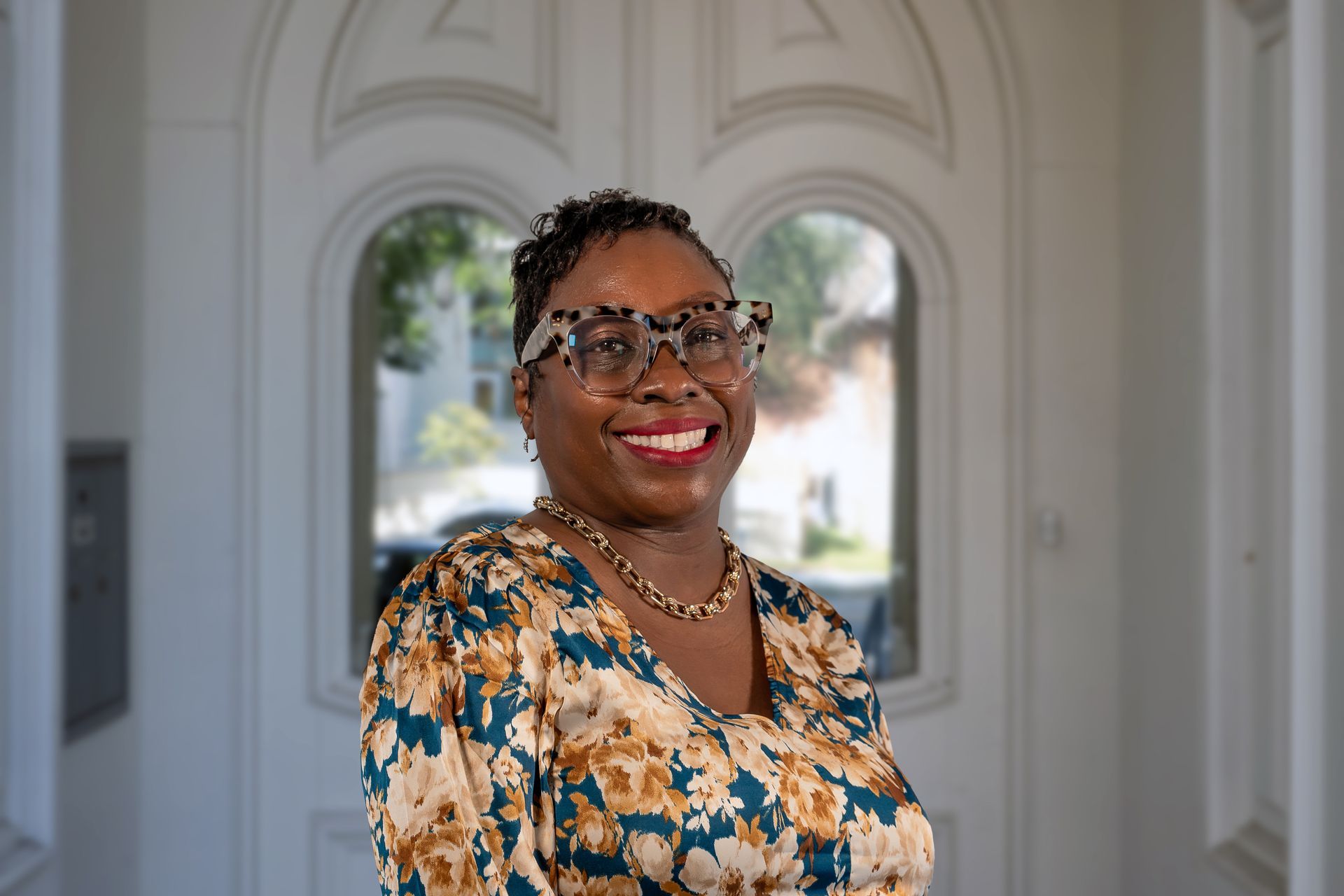 Woman with glasses smiles in front of a white arched doorway. She wears a patterned top and necklace.