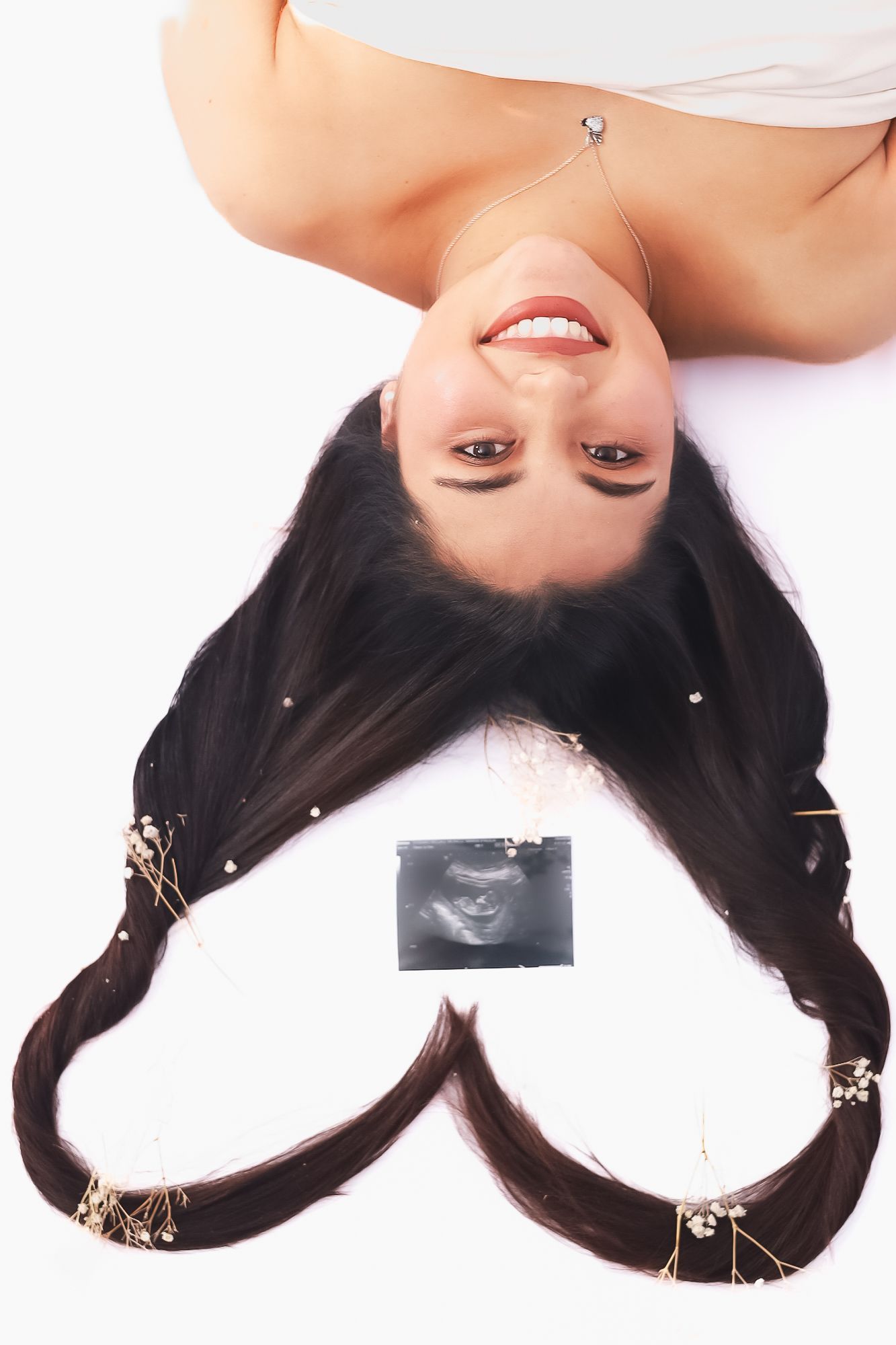 Femme allongée sur fond blanc, les cheveux coiffés en cœur encadrant une image échographique, souriante.