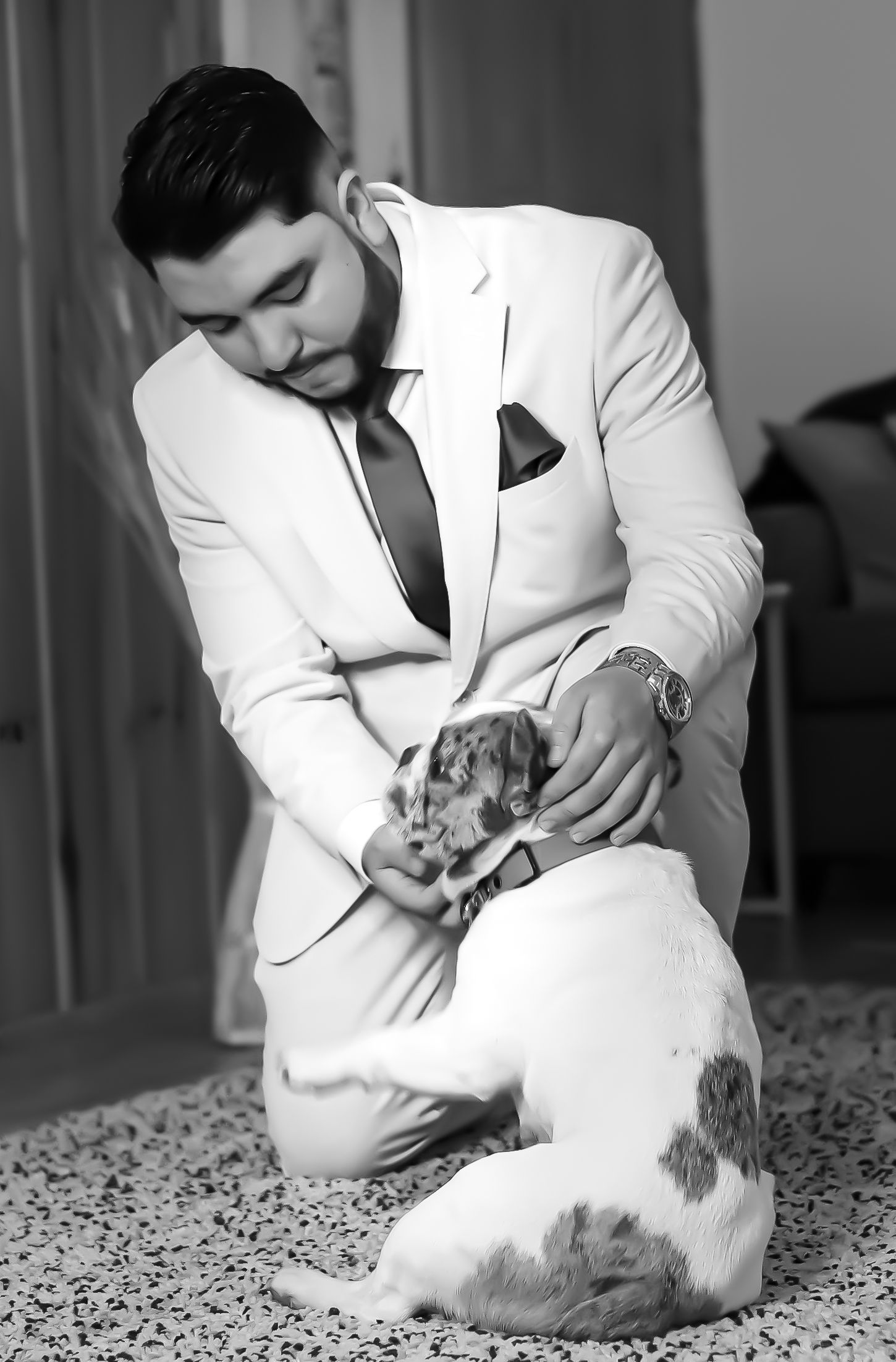 Un homme en costume blanc s'agenouille et caresse un chien tacheté sur un tapis à l'intérieur.