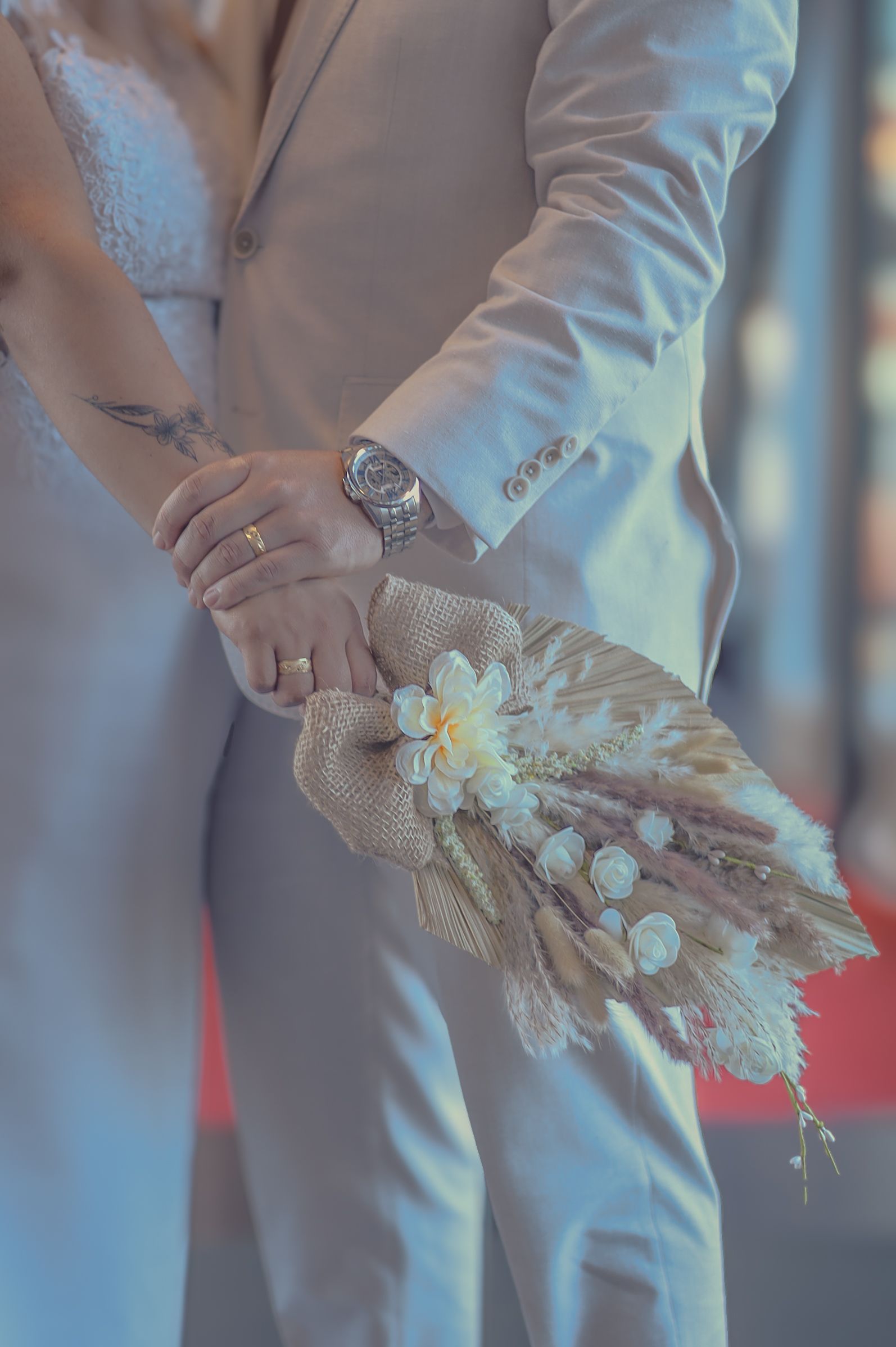 Couple en tenue de mariage, main dans la main et bouquet à la main. Costume beige, robe blanche, alliances visibles.
