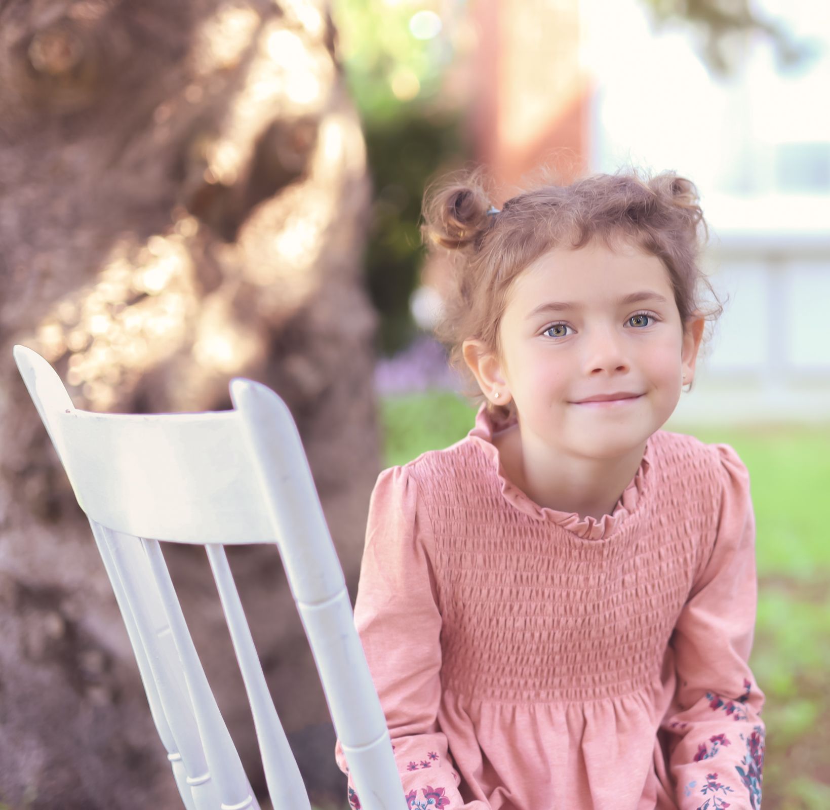 Une fille avec des chignons sourit à côté d'une chaise blanche dans un jardin, vêtue d'une robe rose.