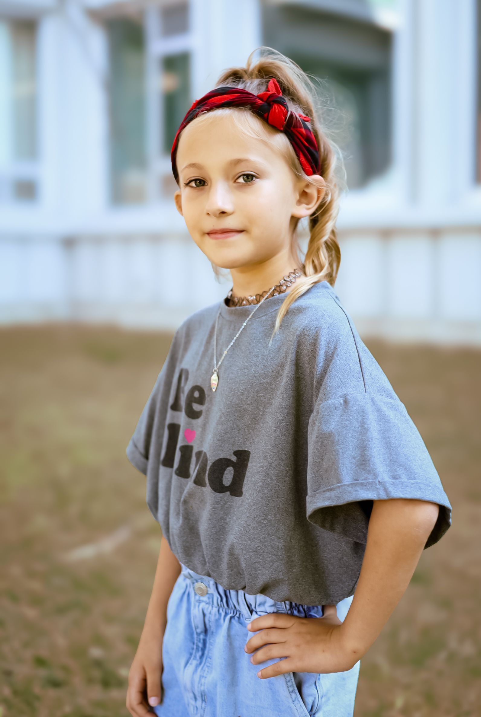 Une fille en t-shirt gris avec l'inscription « Soyez gentils » et un bandeau rouge. Debout dehors, la main sur la hanche.