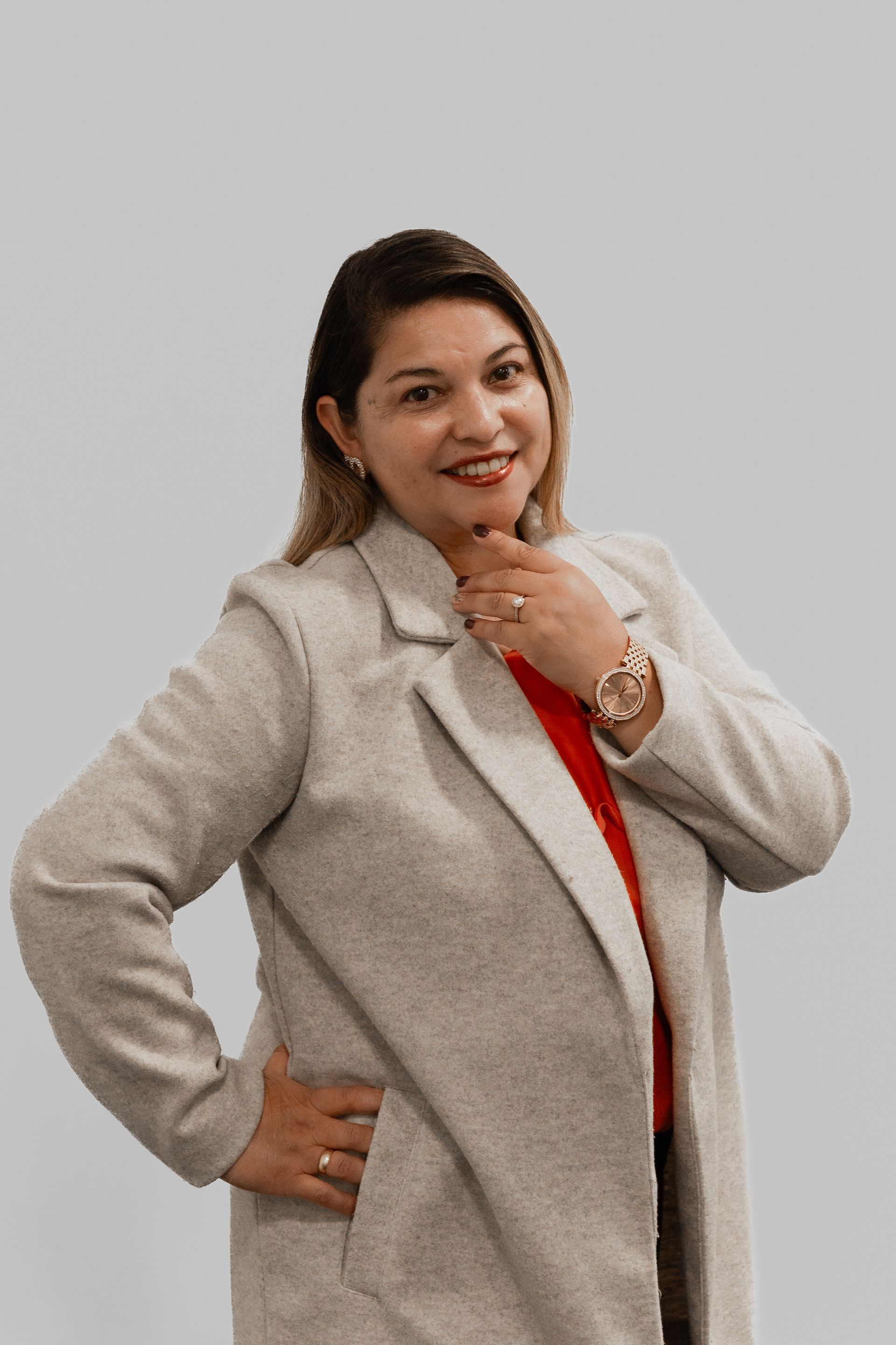 Une femme portant un manteau gris et une chemise rouge est debout, les mains sur les hanches.