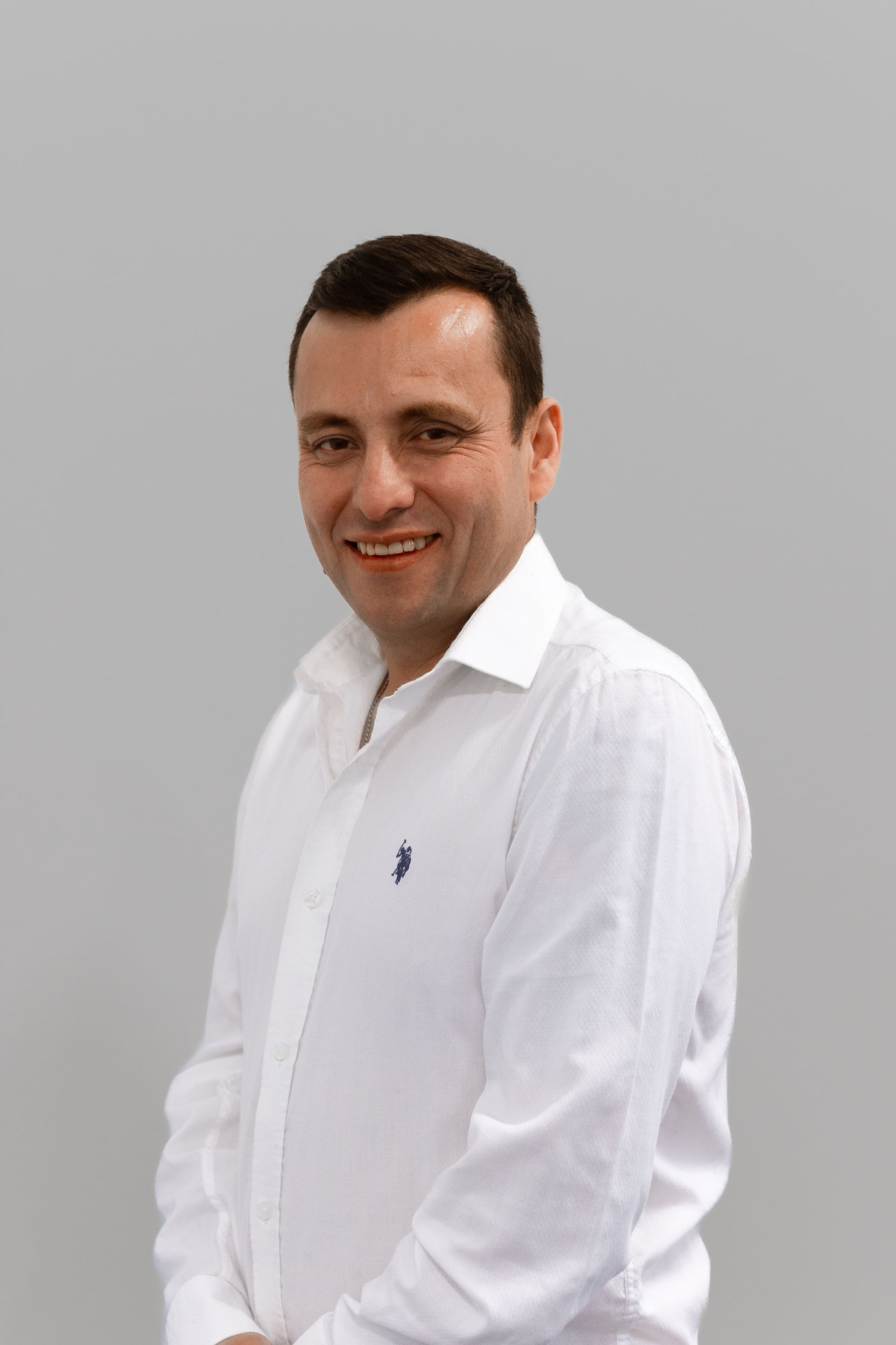 Un homme en chemise blanche sourit et se tient devant un fond gris.