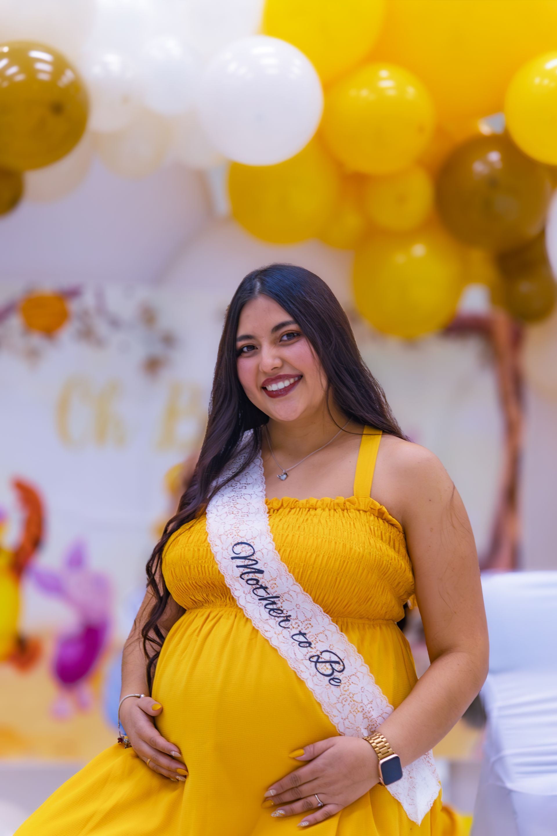 Femme enceinte en robe jaune avec une écharpe « Future Maman » souriant à une baby shower. Ballons en arrière-plan.