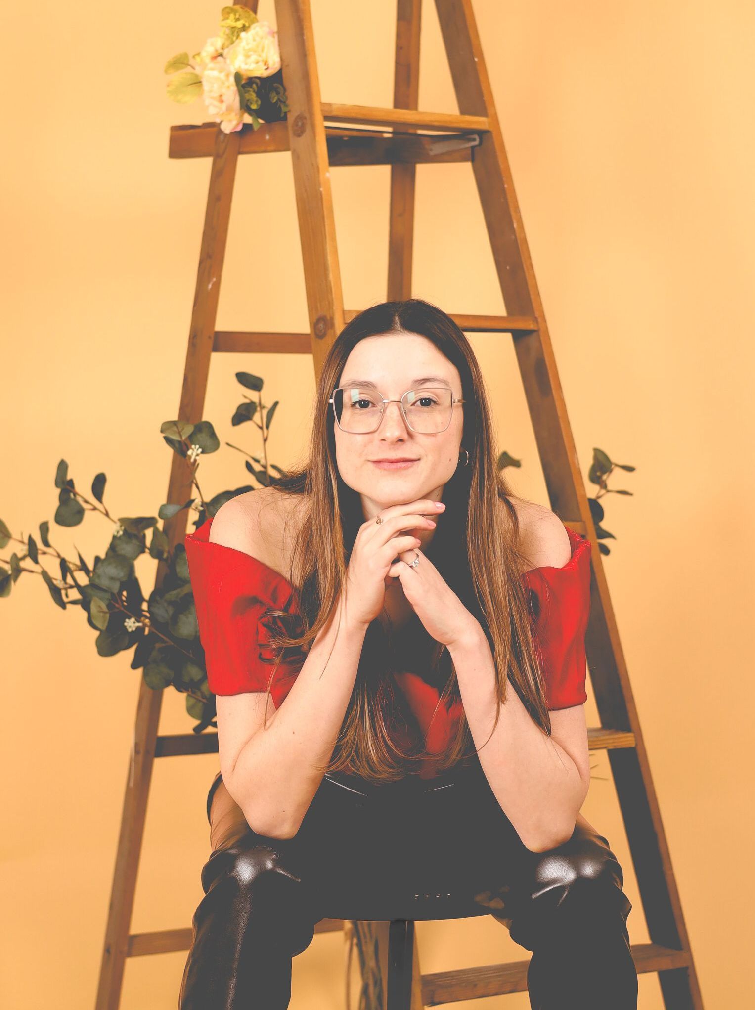 Femme portant des lunettes et un haut rouge, assise sur une échelle sur fond beige avec des fleurs et de la verdure.