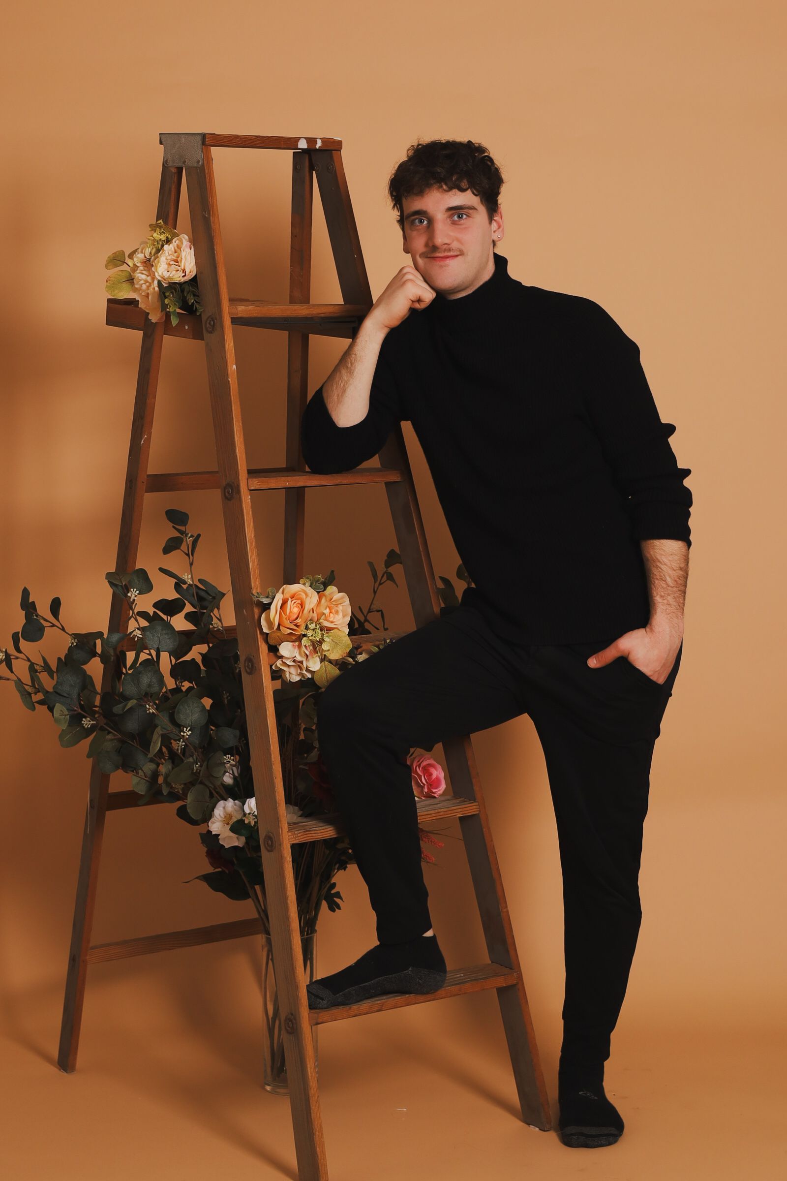 Un homme en col roulé noir s'appuie contre une échelle en bois décorée de fleurs.