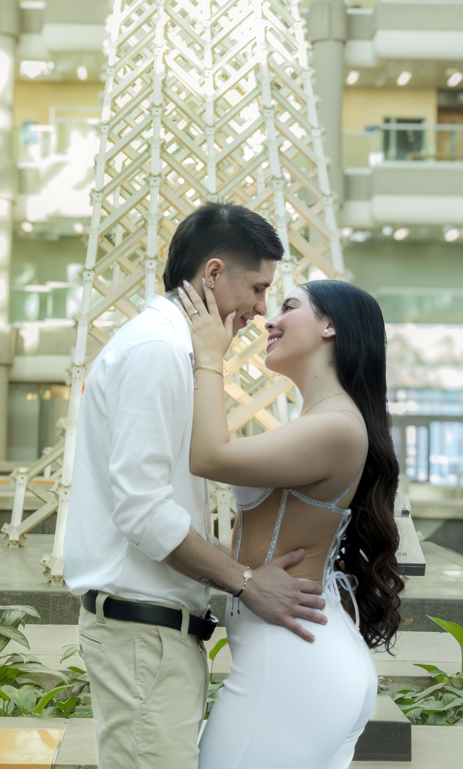 Couple enlacé ; homme en chemise blanche et pantalon kaki, femme en robe blanche, près d'une structure décorative.