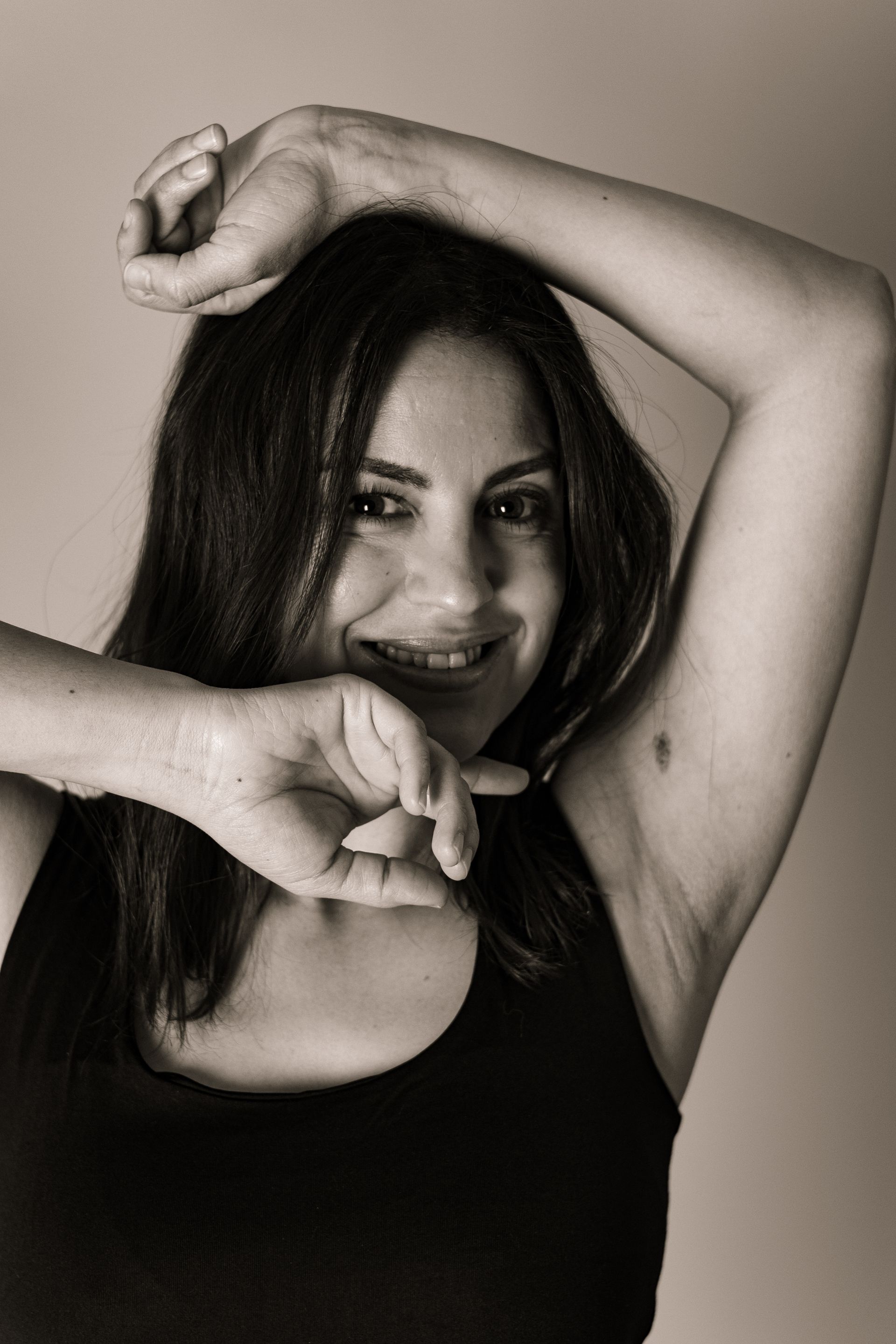 Femme souriante, les bras levés, une main près du visage, portant un débardeur noir.