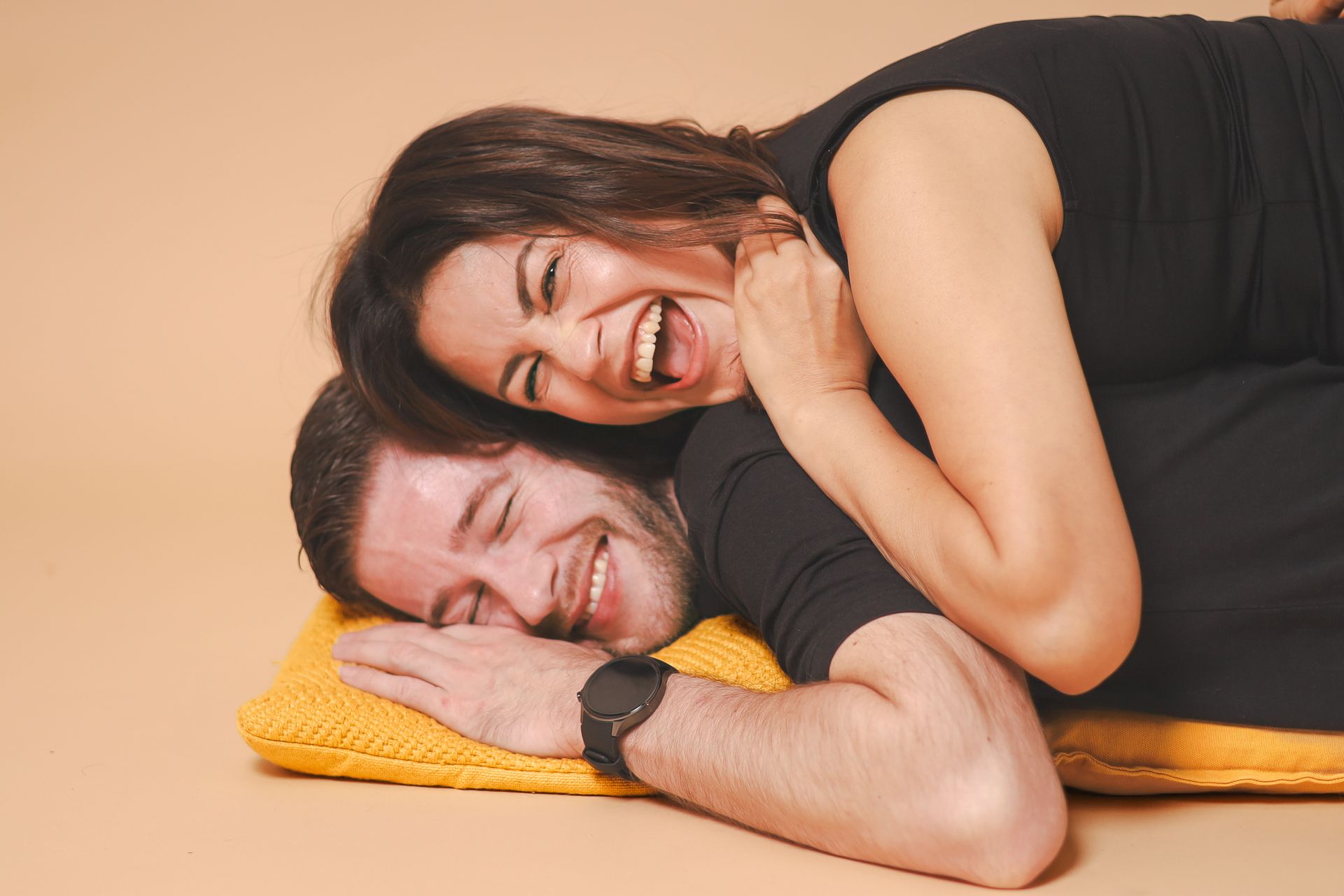 Une femme rit joyeusement sur un homme allongé sur un oreiller jaune, tous deux en chemise noire, sur un fond beige.