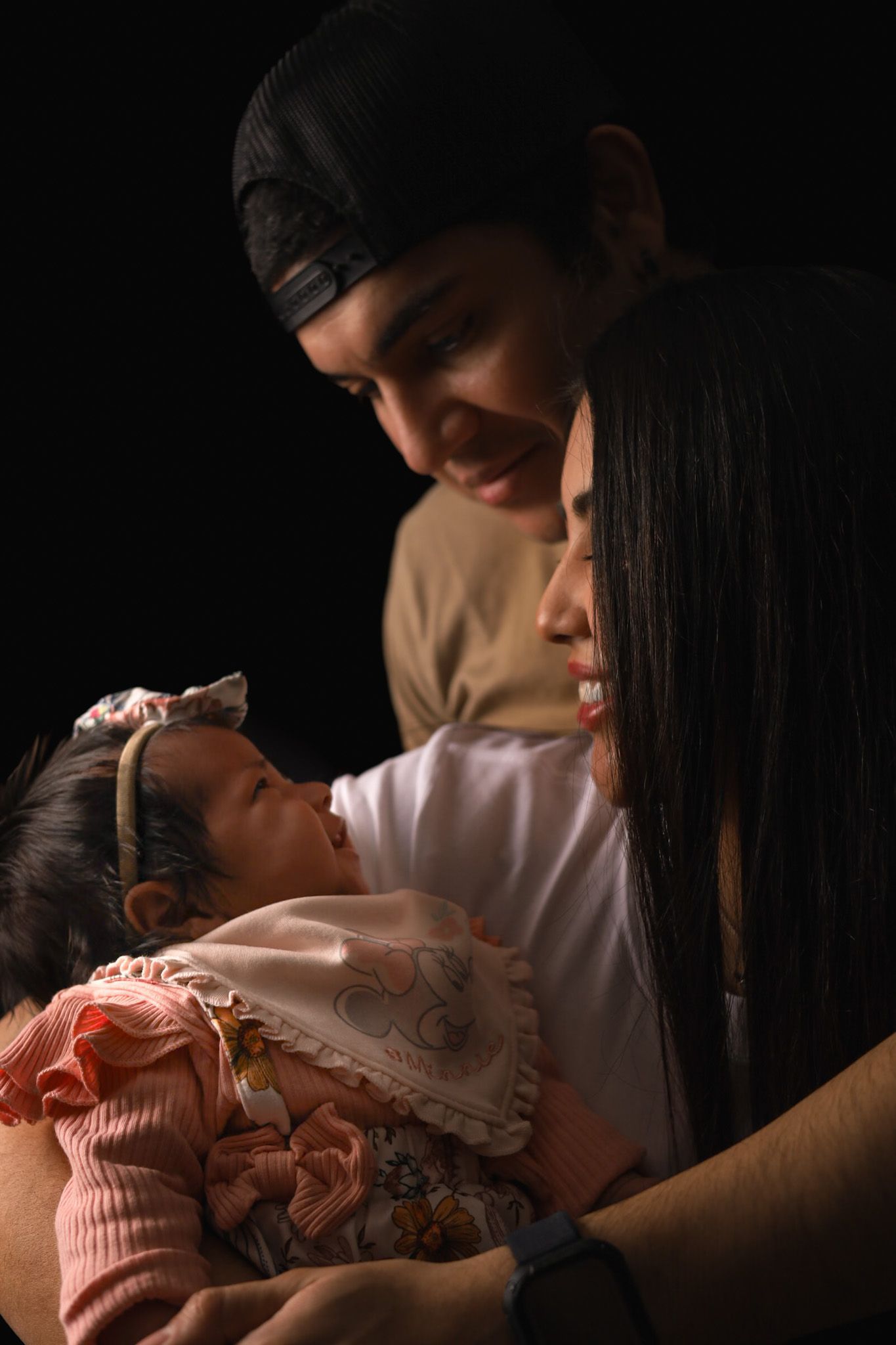 Des parents regardent avec adoration un bébé dans leurs bras, un fond sombre.
