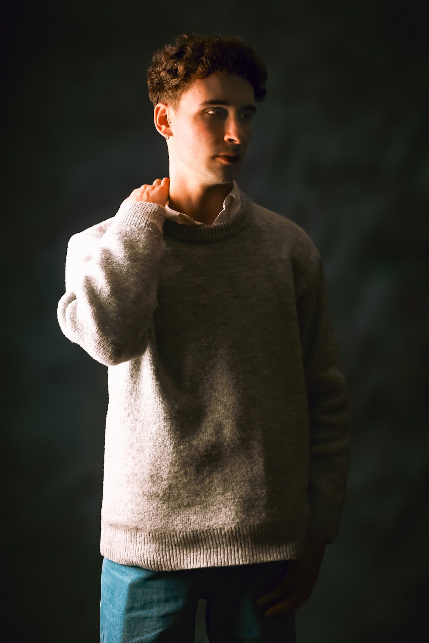 Homme en pull gris, main droite sur le cou, regardant vers la gauche sur un fond sombre.