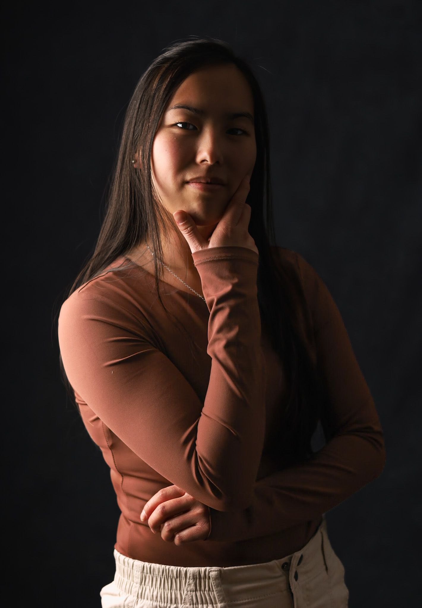 Femme en chemise marron à manches longues avec sa main sur son menton, sur un fond sombre.