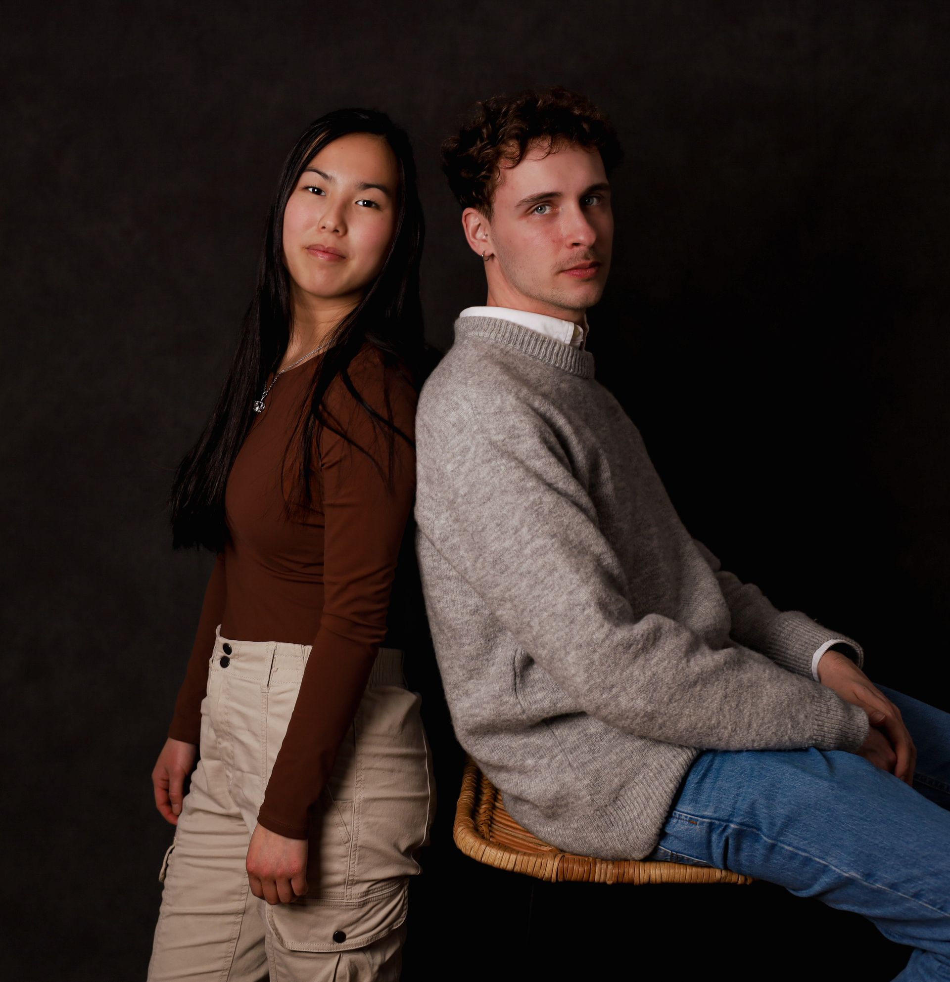 Deux personnes sont assises dos à dos, face à face. Fond sombre. Femme : haut marron, pantalon beige. Homme : pull gris, jean.