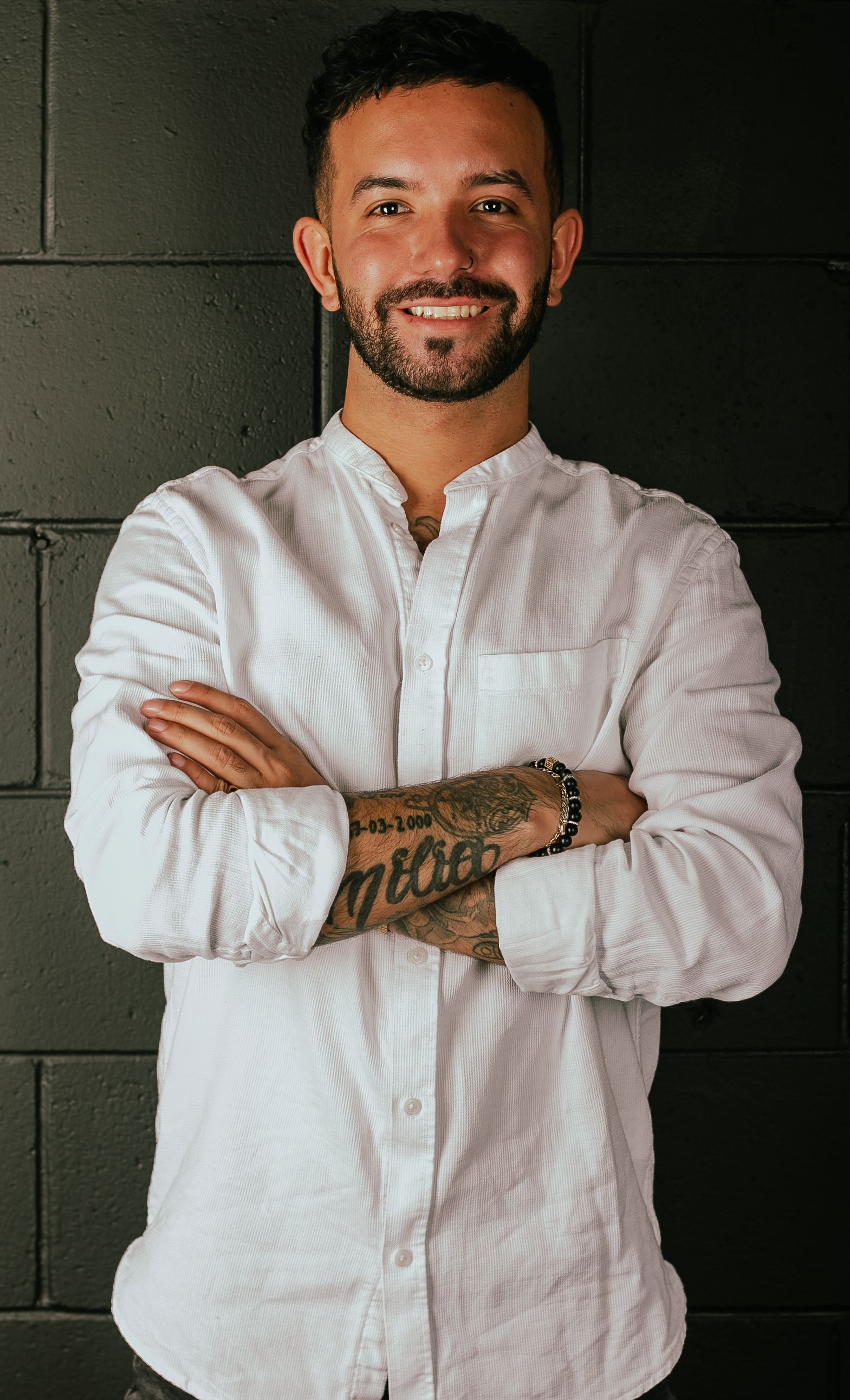 Homme aux bras croisés, souriant, portant une chemise blanche ; mur de briques sombres en arrière-plan.