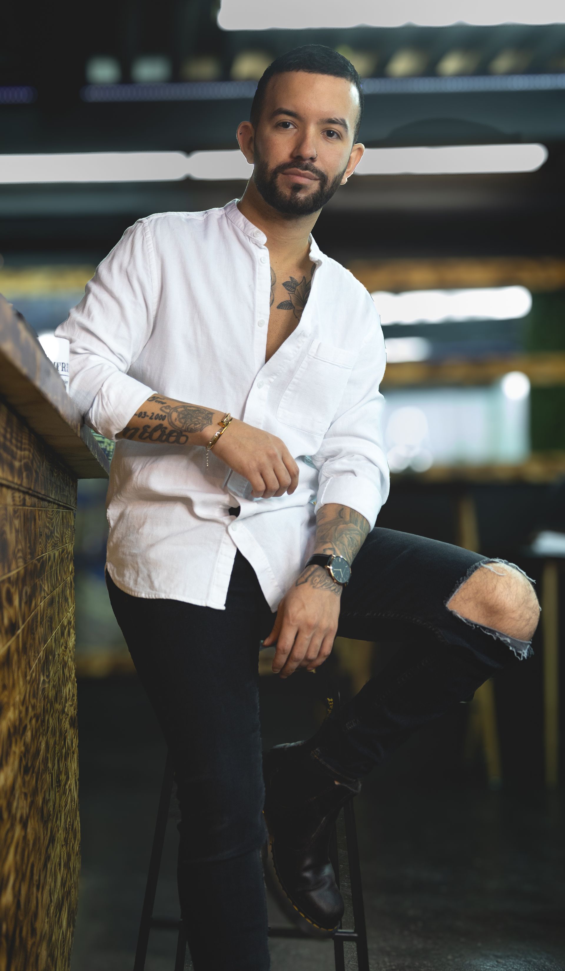 Un homme avec des tatouages, une chemise blanche et un jean noir, s'appuie sur le bar et regarde la caméra.