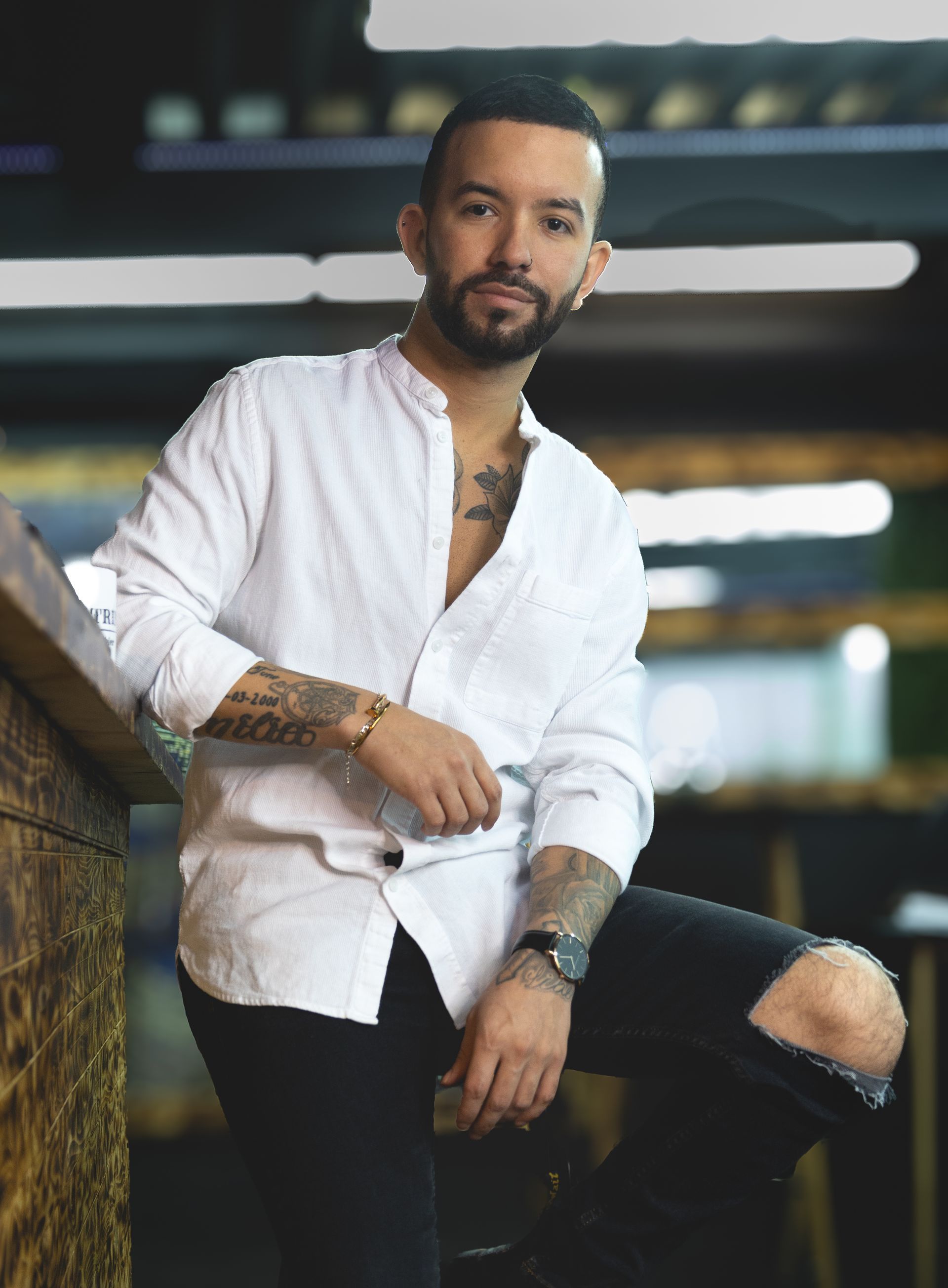 Un homme avec une barbe est assis sur un tabouret de bar portant une chemise blanche et un jean noir.