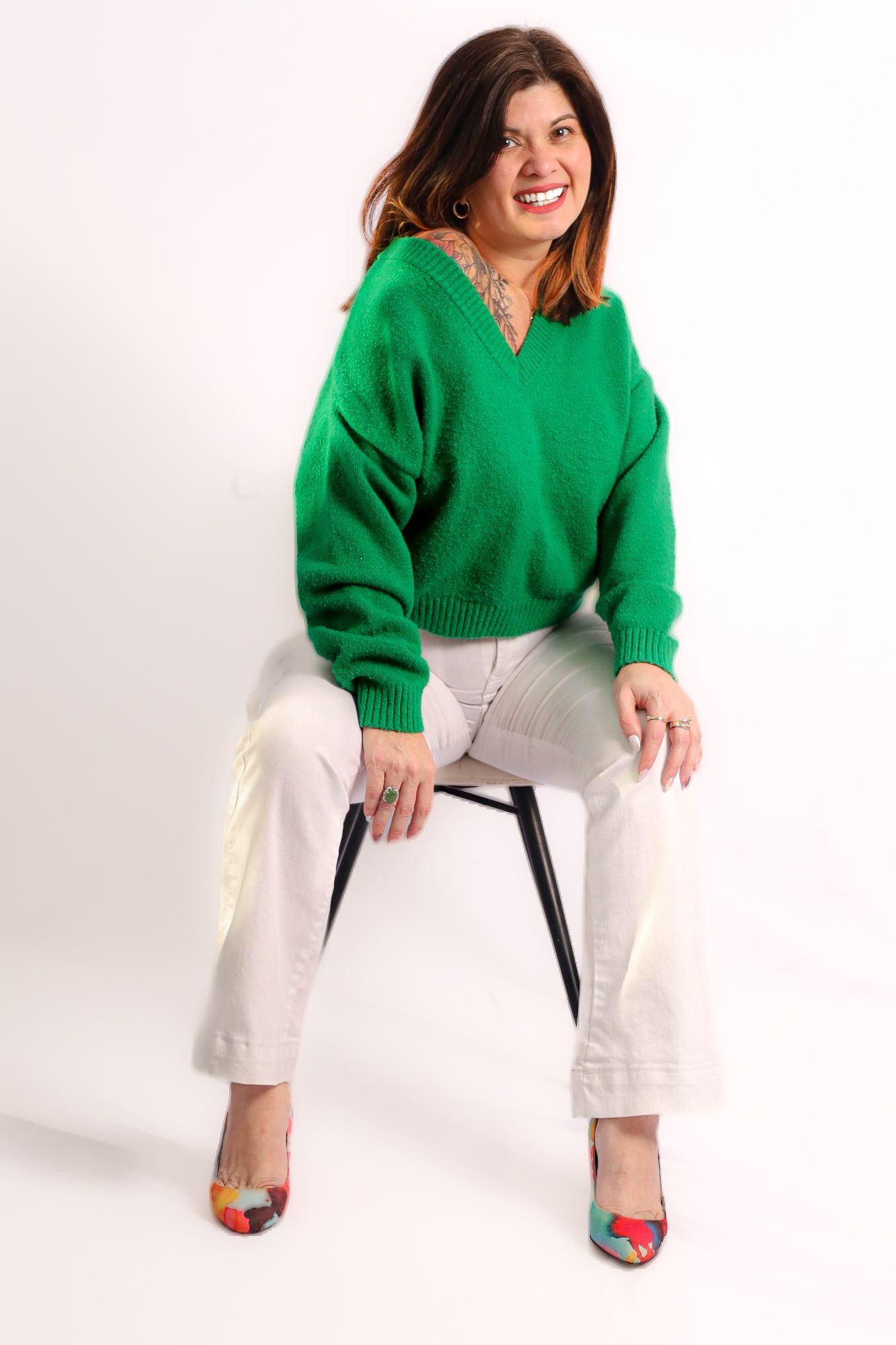 Une femme en pull vert et pantalon blanc est assise en souriant sur un tabouret noir.