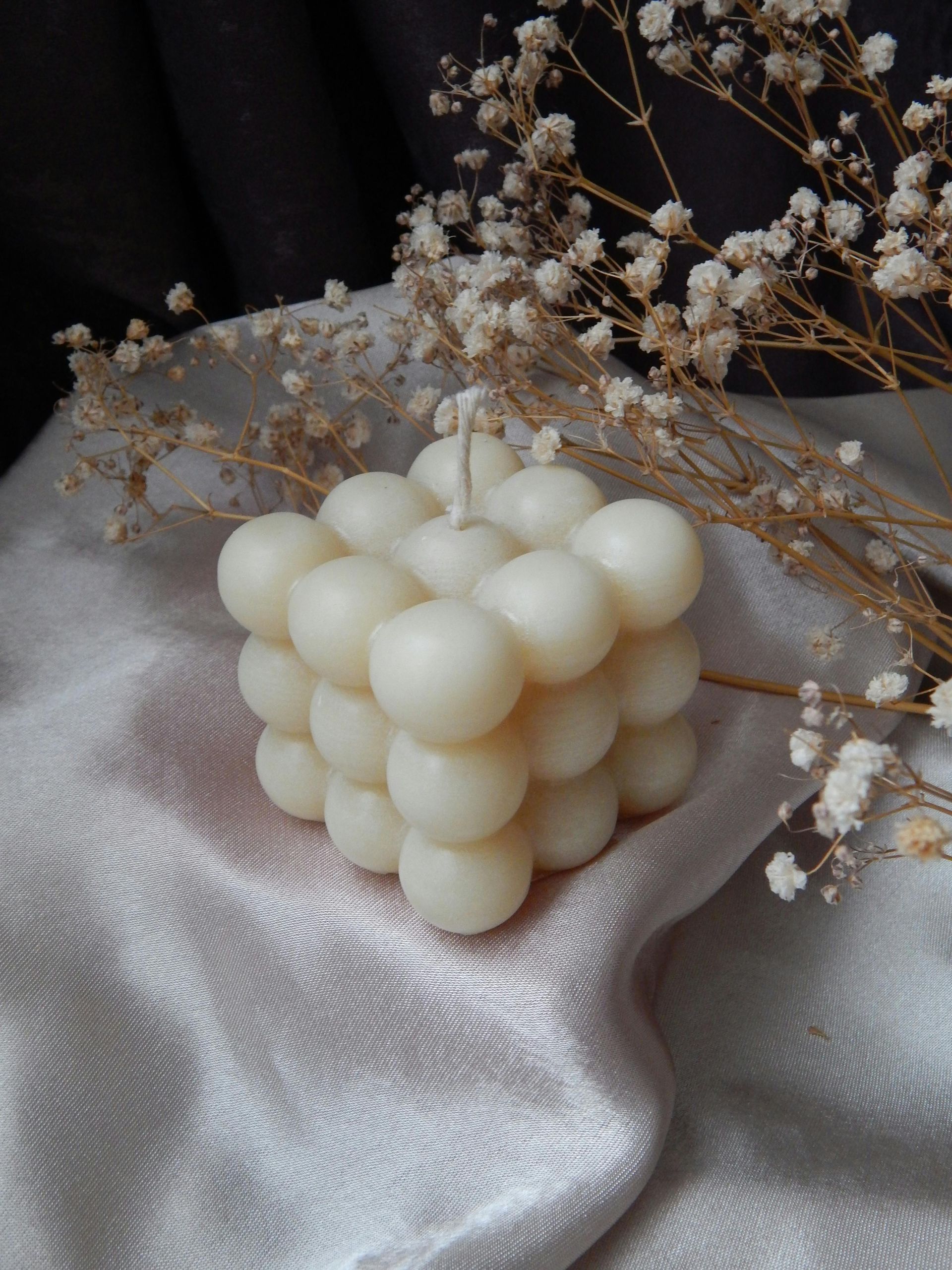 Une bougie blanche en forme de cube est posée sur un tissu blanc à côté de fleurs séchées.