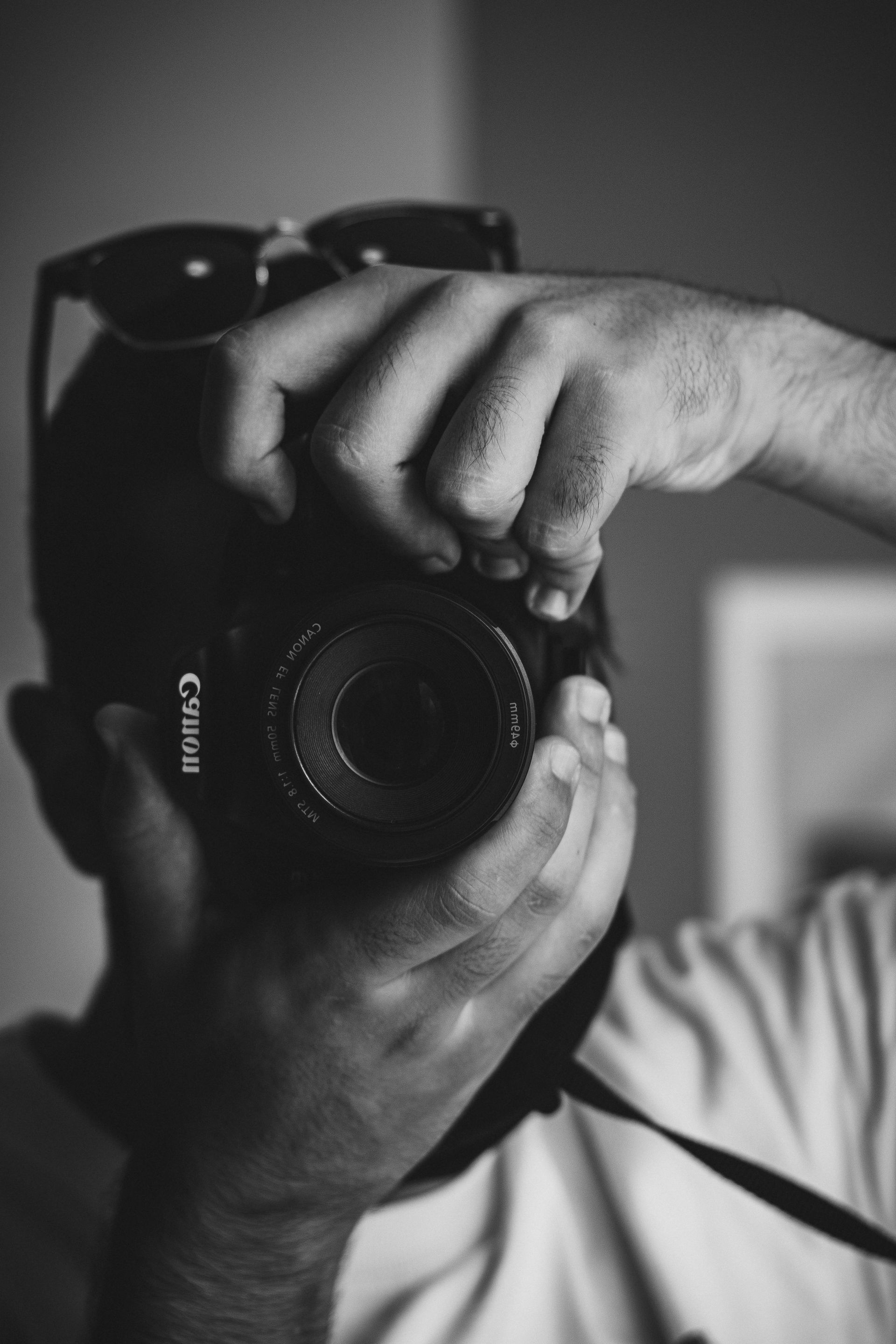 Gros plan en noir et blanc d'une personne tenant un appareil photo reflex numérique Canon devant son visage, des lunettes de soleil posées sur sa tête.