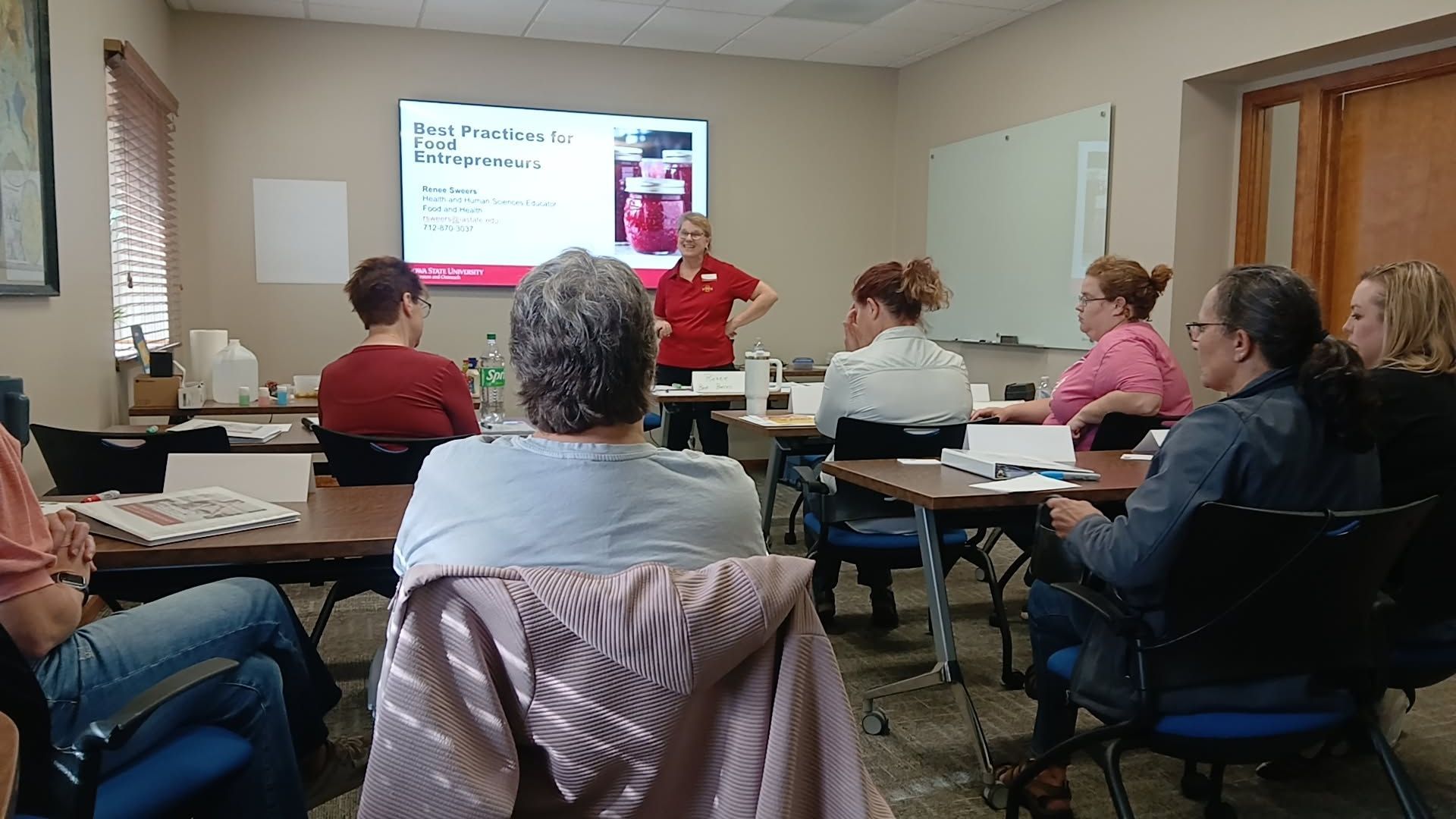 A woman in a red shirt presents in a classroom. Attendees sit at desks, looking at a screen.