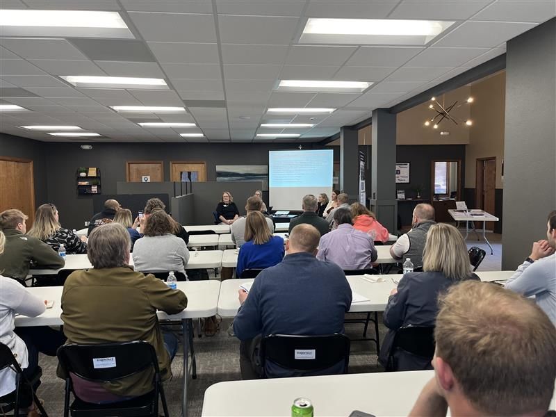 People seated at tables in a conference room, facing a screen. A person is presenting.