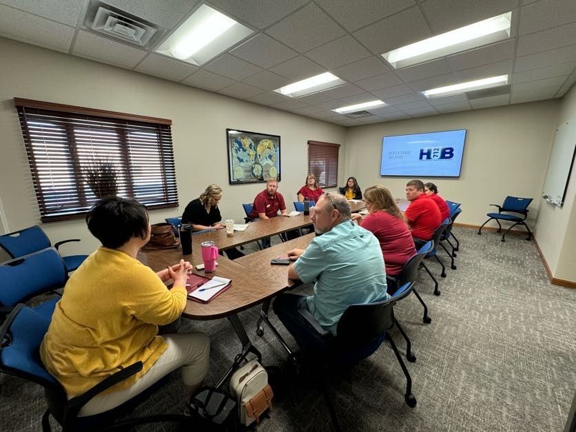 People seated around a conference table in a meeting room, discussing. A screen shows "HUB" logo.