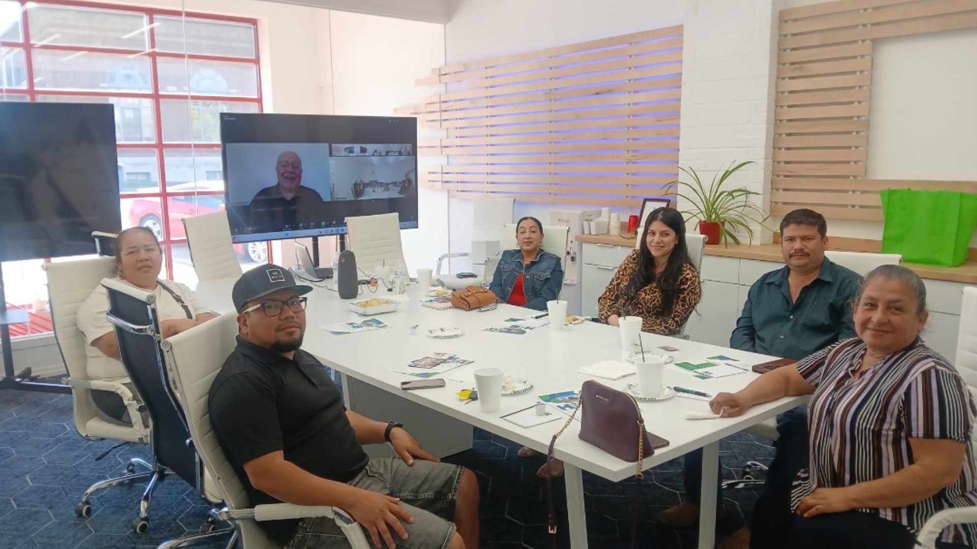 People seated around a table with a screen showing a person on a video call in a bright office.