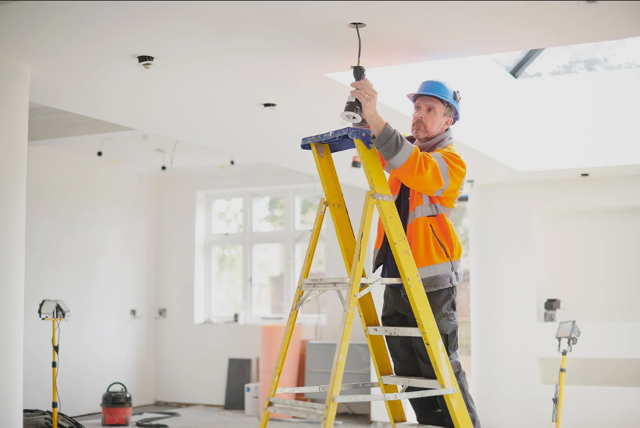 Electrician on a yellow ladder installing a light fixture in a white room; wearing a hard hat and safety vest.