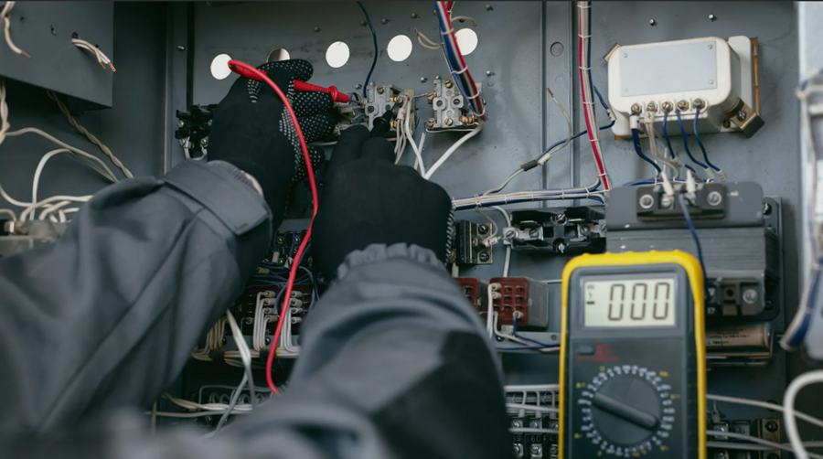Electrician in black gloves using a multimeter to test wires in a control panel.