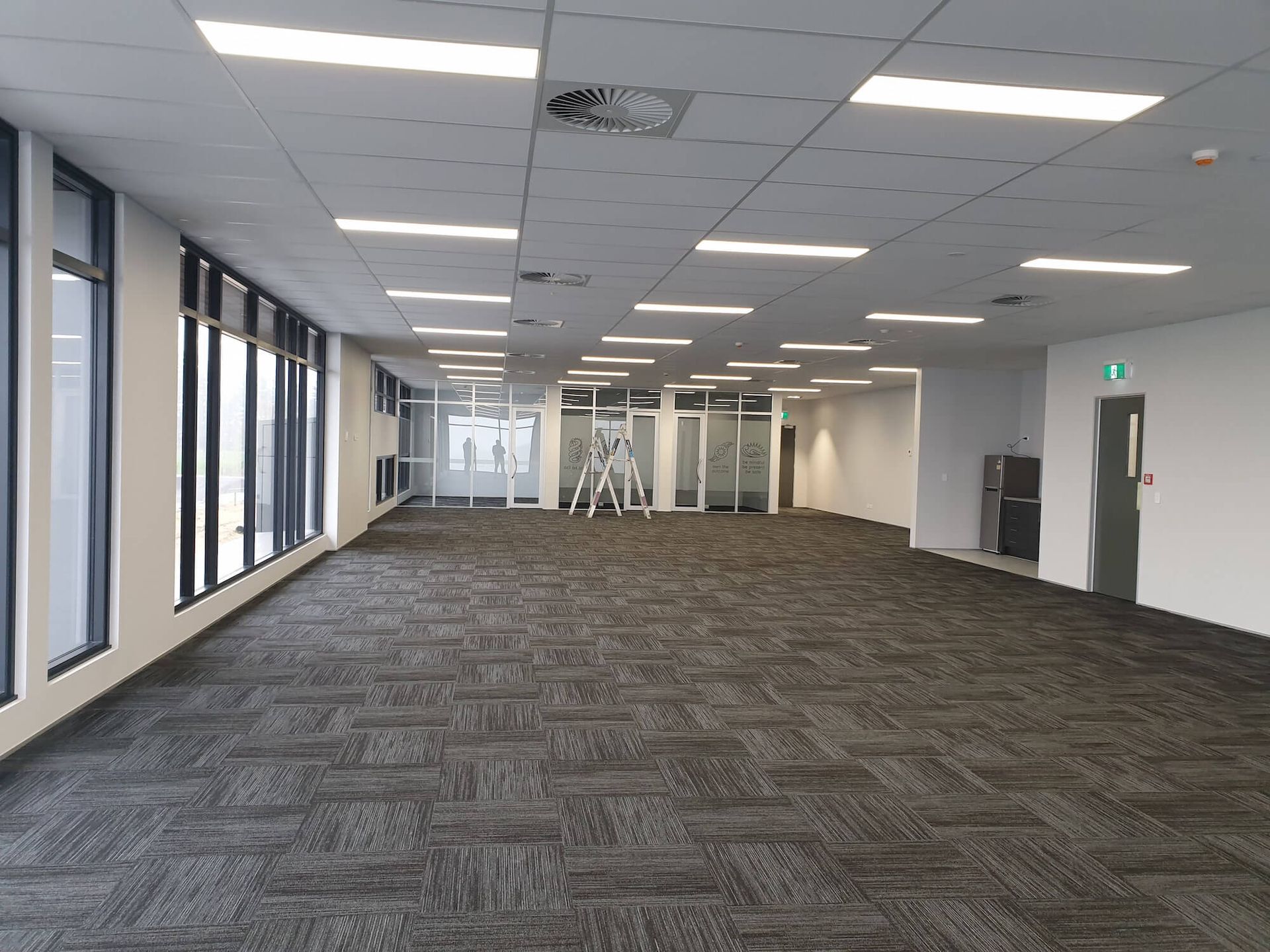 Empty office space with large windows, carpet, and overhead lights.