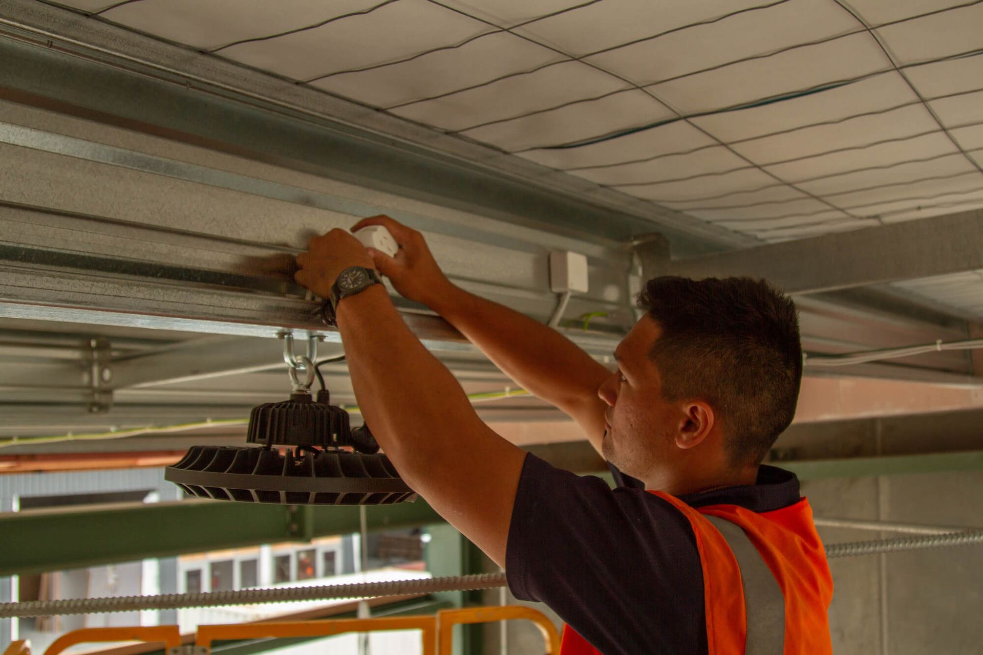 Man in orange vest installing a large overhead light fixture in a commercial building. His hands work above the ceiling.