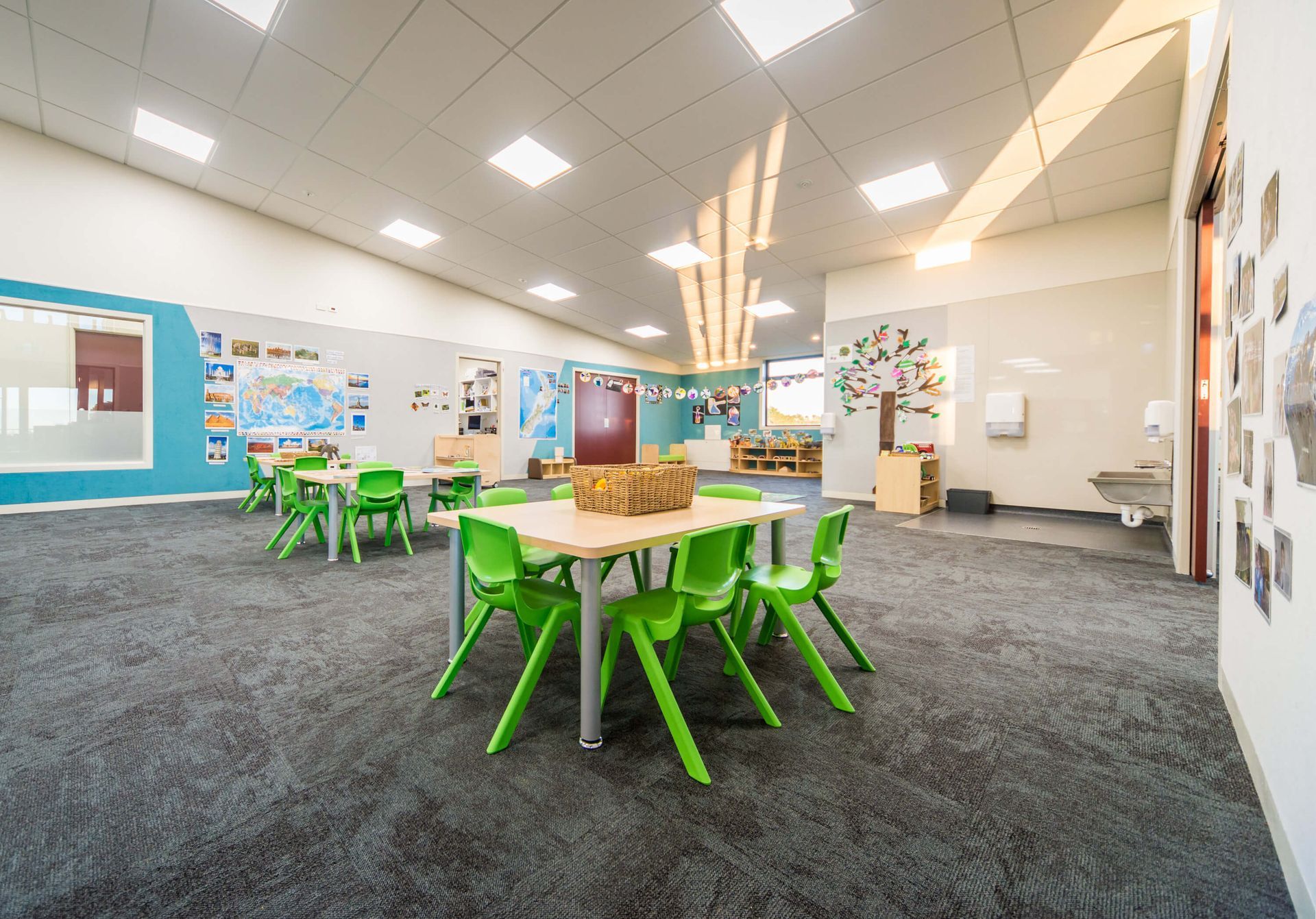 Bright, airy preschool classroom with green chairs around tables on grey carpet. Decorated walls with educational displays.