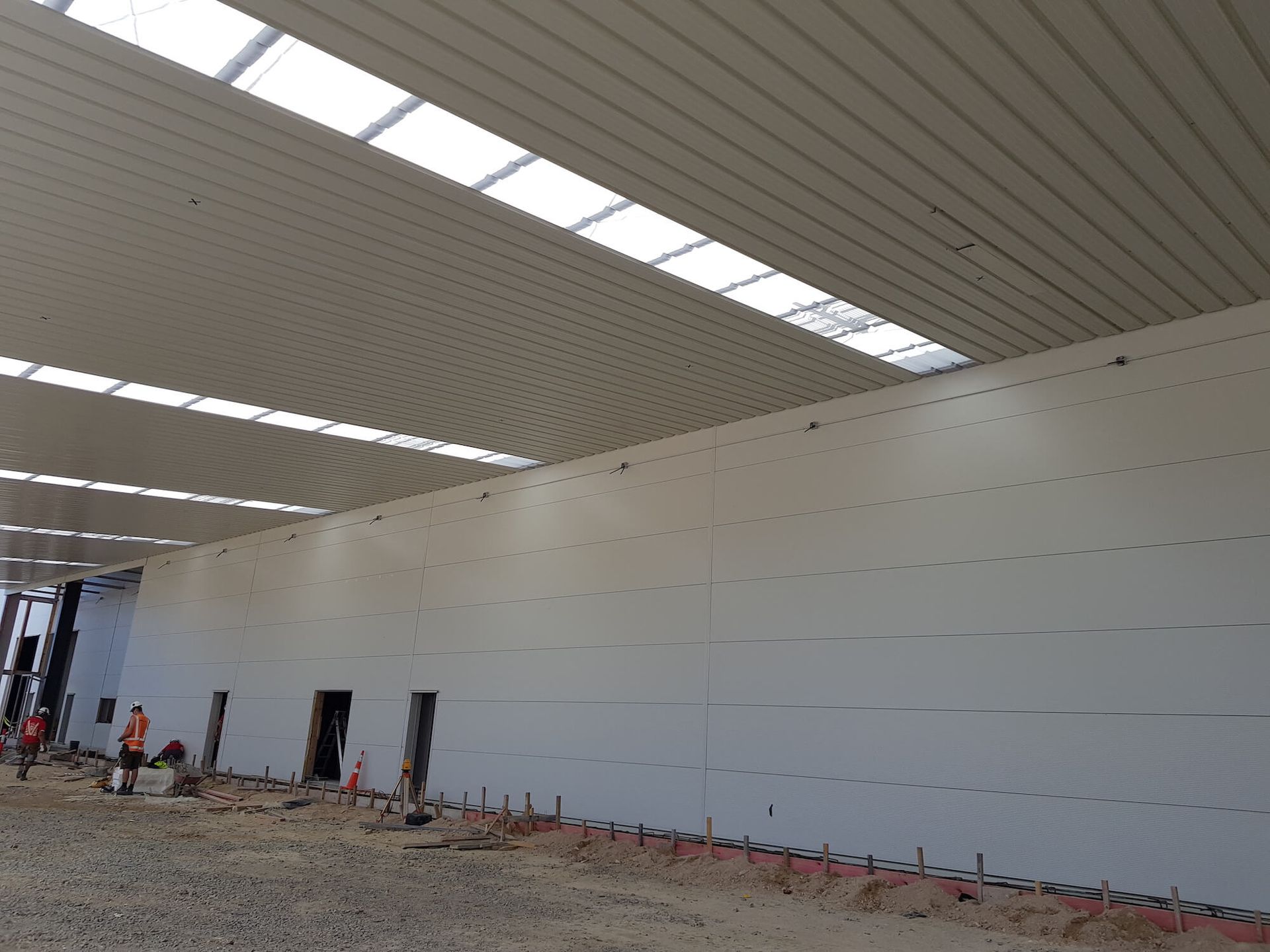 Interior view of a large industrial building under construction with white walls and ceiling panels, skylights, and workers present.