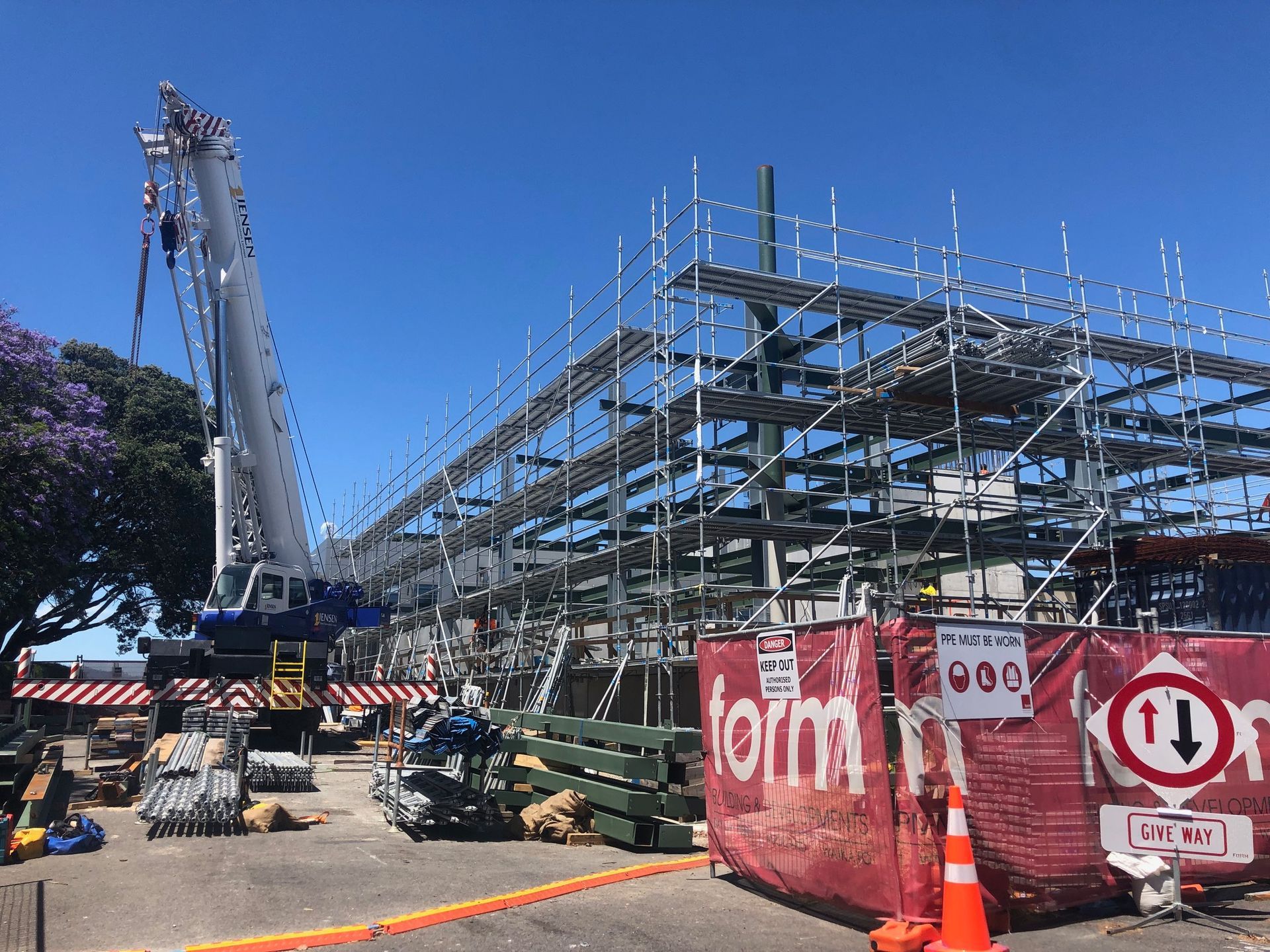 Construction site with a crane and scaffolding under a bright blue sky. Red barriers and traffic cones are set up.