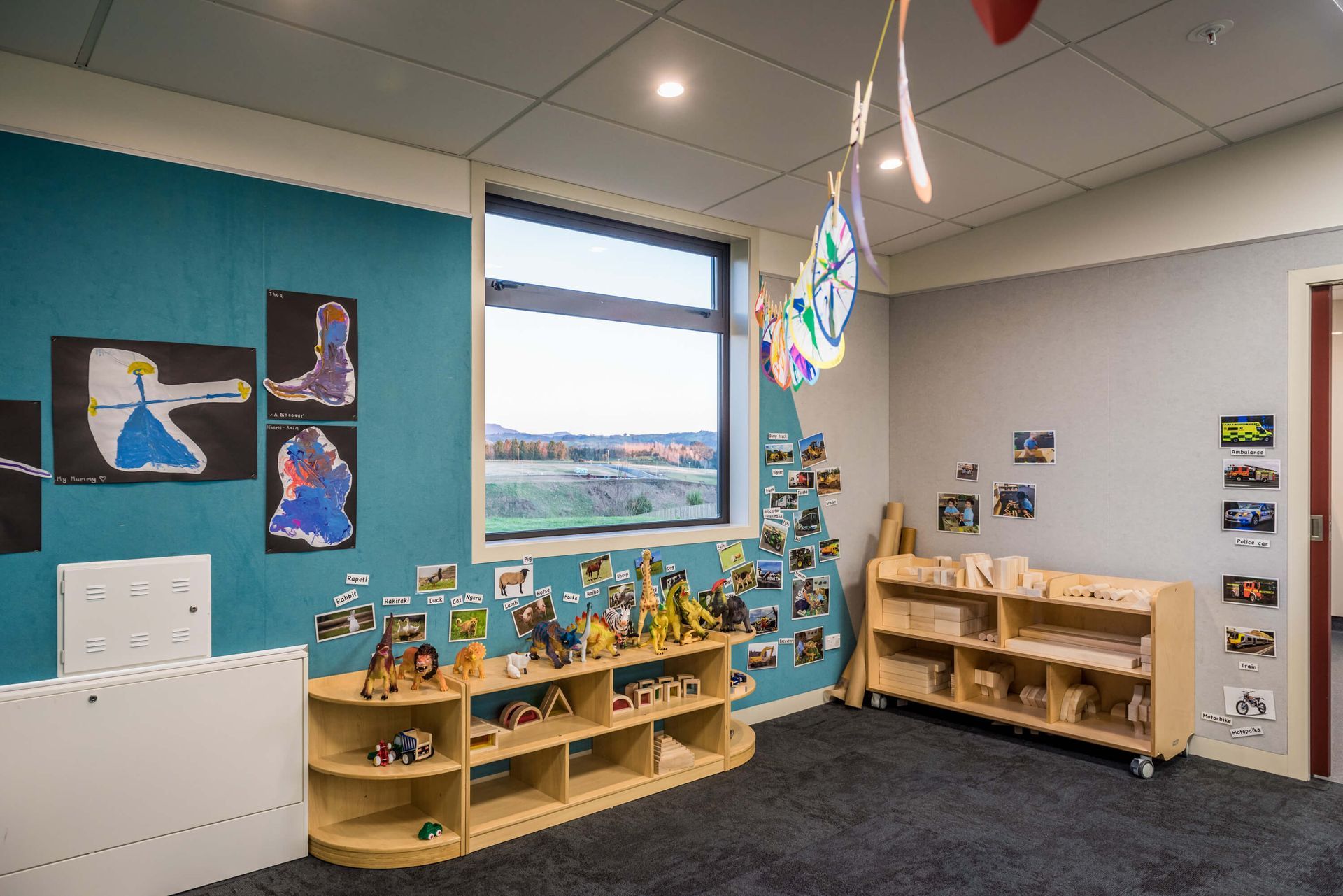 Childcare room with turquoise and gray walls, open shelves with toys, artwork, and a large window with an outdoor view.