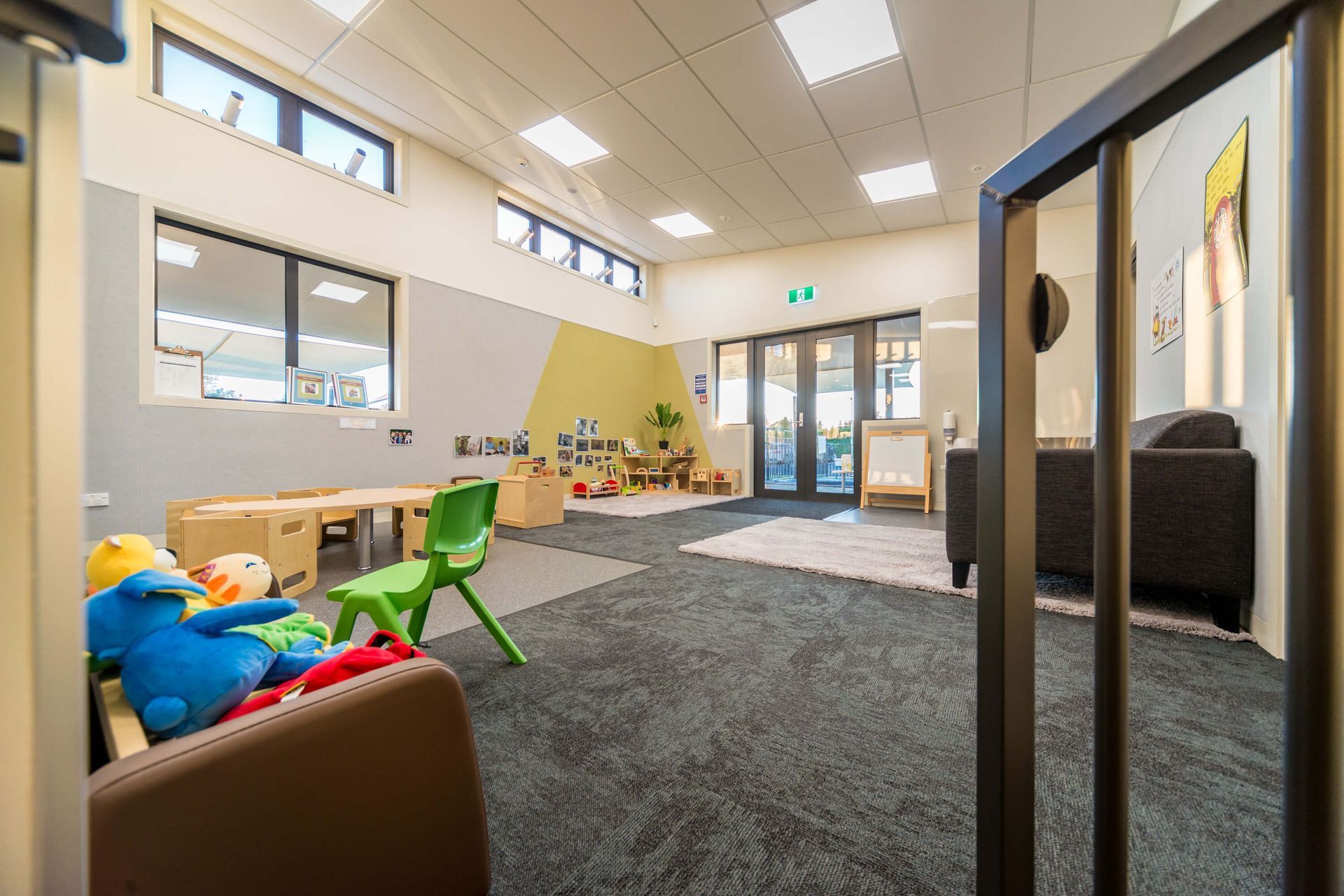Childcare room with tables, chairs, toys, and an open doorway to an outdoor area. Green and yellow accents on the walls.