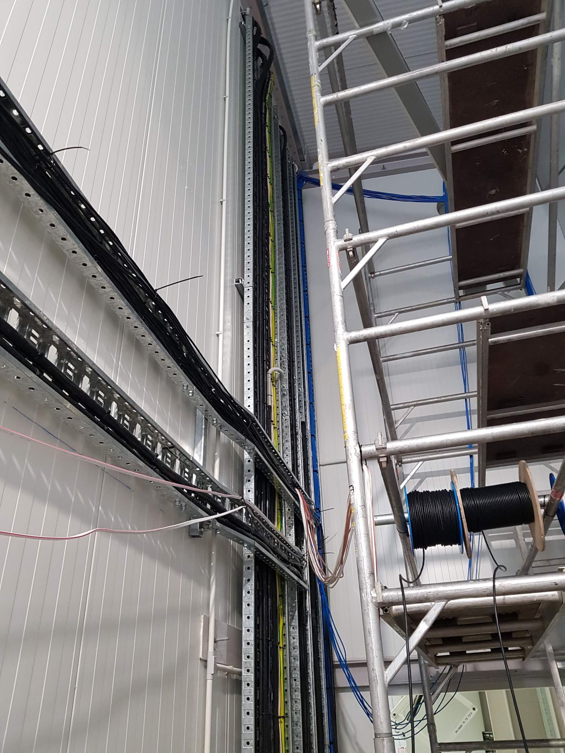 Cables running up a wall, secured in trays. A scaffolding is set up next to the wall with wire spools at the base.