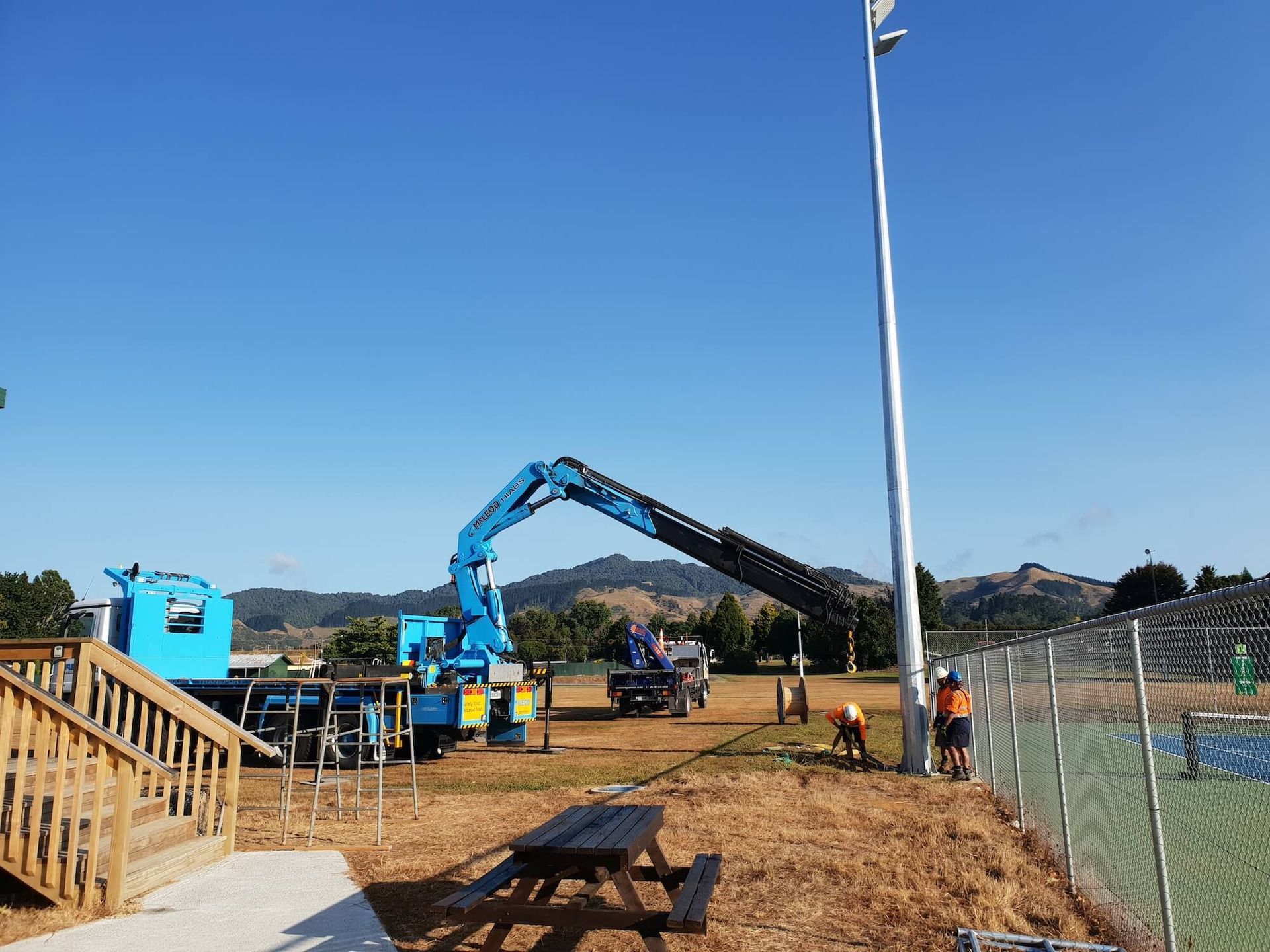 A blue crane lifting a tall metal light pole near a sports field with workers, a fence, and a picnic table under a clear blue sky.