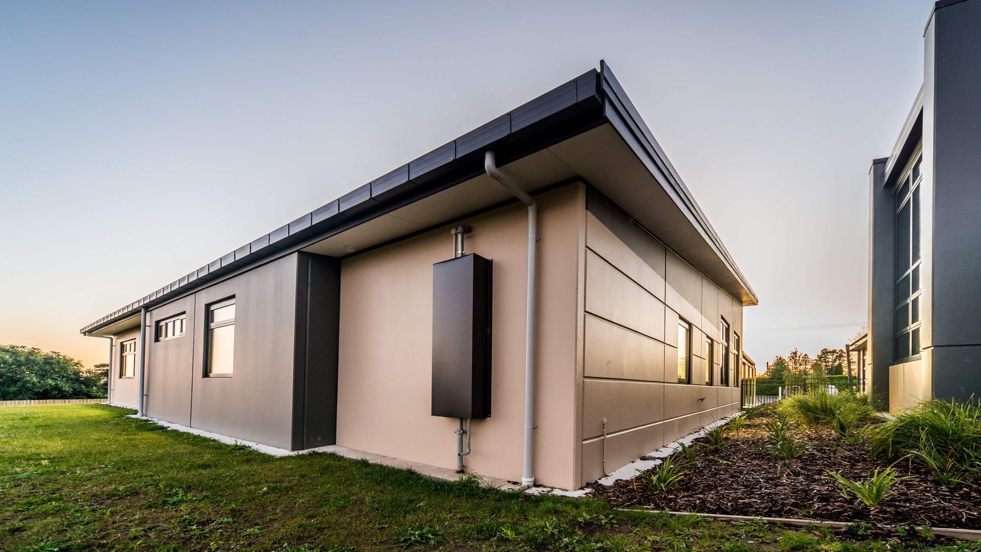 Modern industrial building with gray and beige panels, black roof, and small windows, set in a grassy area.