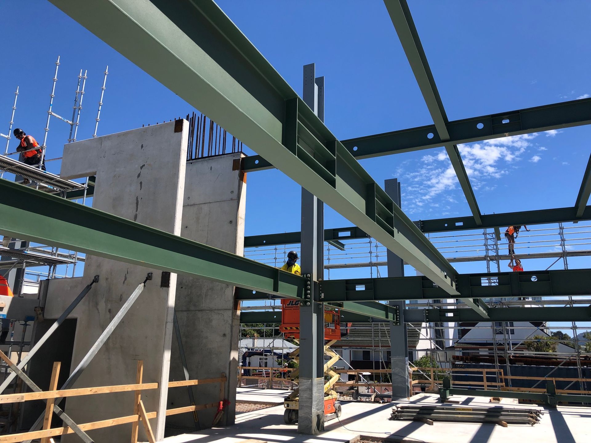 Construction site with steel beams and concrete columns under a blue sky. Workers are on scaffolding, assembling the building frame.